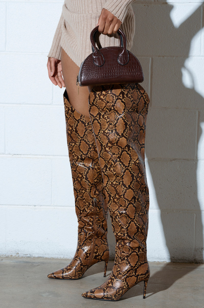 A person in AZALEA WANG TARRANT SNAKE WESTERN TOPLINE boots and a beige ribbed sweater dress holds a small brown handbag, standing confidently against a white wall.