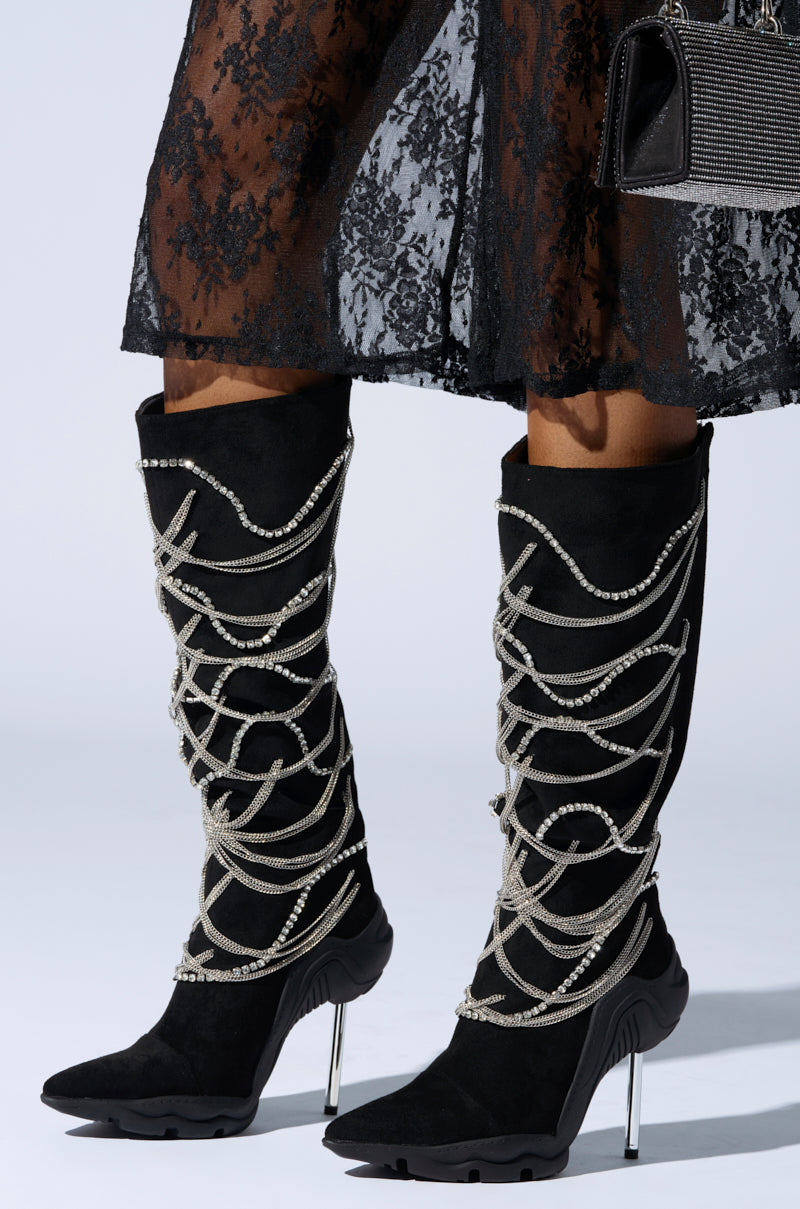 The AZALEA WANG TATY Black Chain Embellished Boot features suede uppers adorned with silver chains. The stiletto heels add a sleek touch when worn with a sheer lace skirt and paired with a small, glittery black handbag.