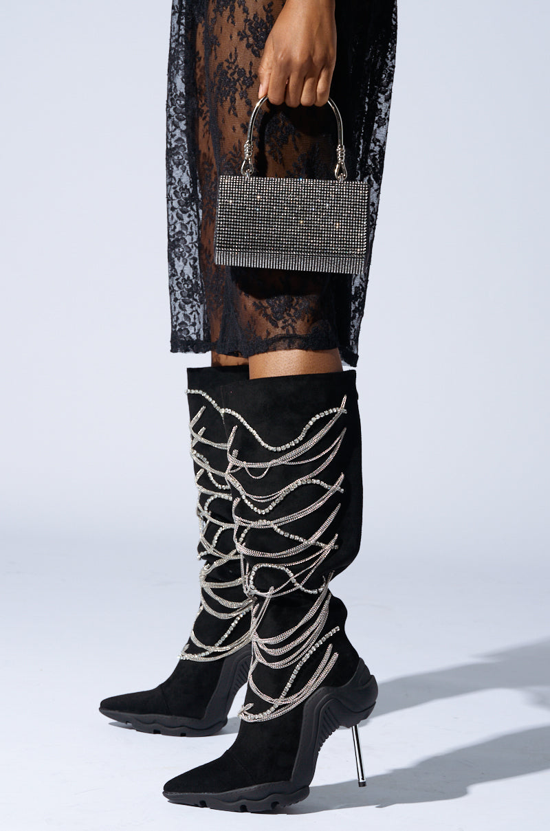 Wearing the AZALEA WANG TATY BLACK CHAIN EMBELLISHED BOOTS, a person complements a sheer lace skirt while holding a small, studded handbag on a plain white background.