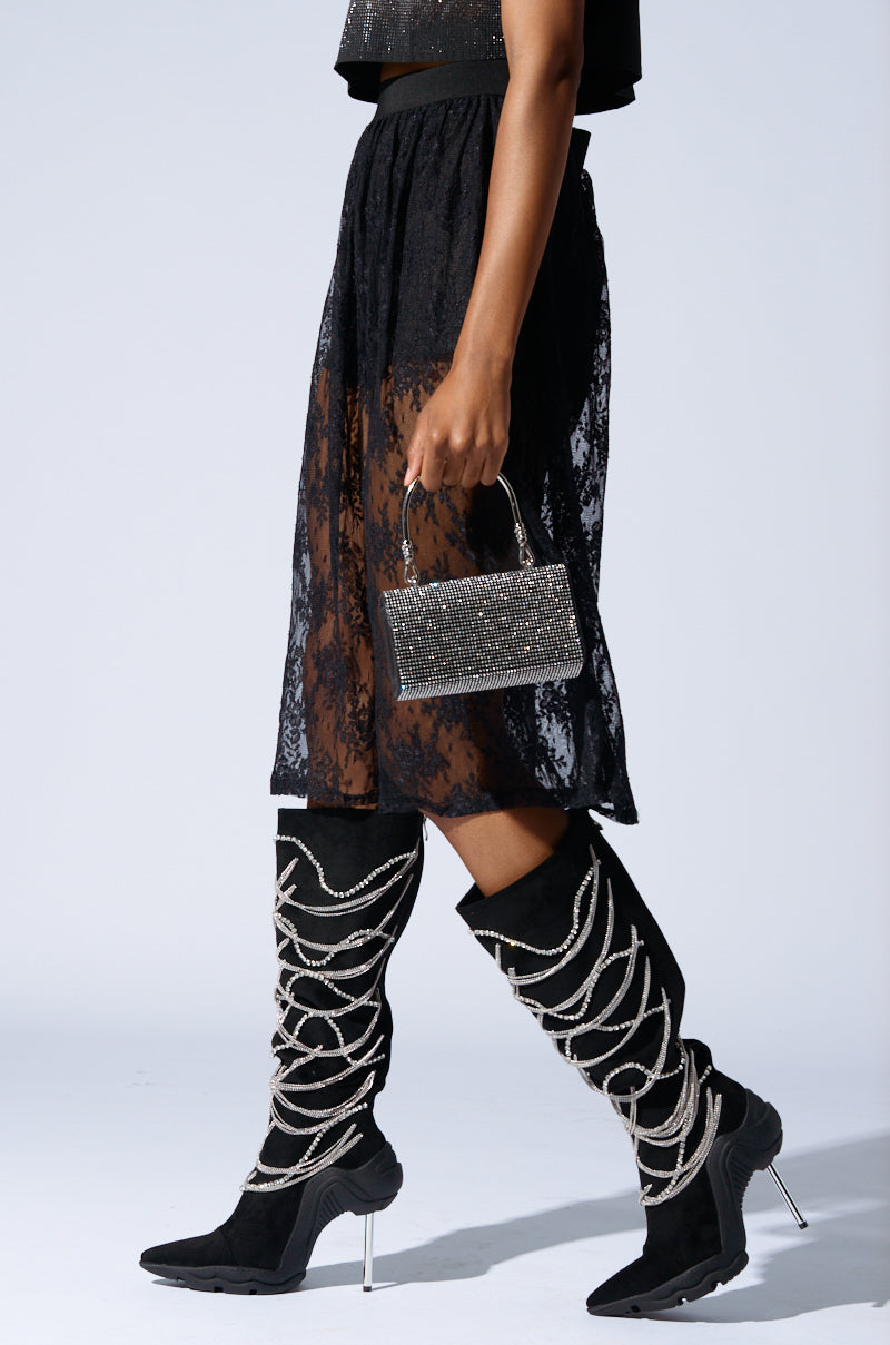 A person wearing a black lace skirt, black top, and the AZALEA WANG TATY BLACK CHAIN EMBELLISHED BOOT with a stiletto heel is holding a small, glittery silver handbag against a light gray background.