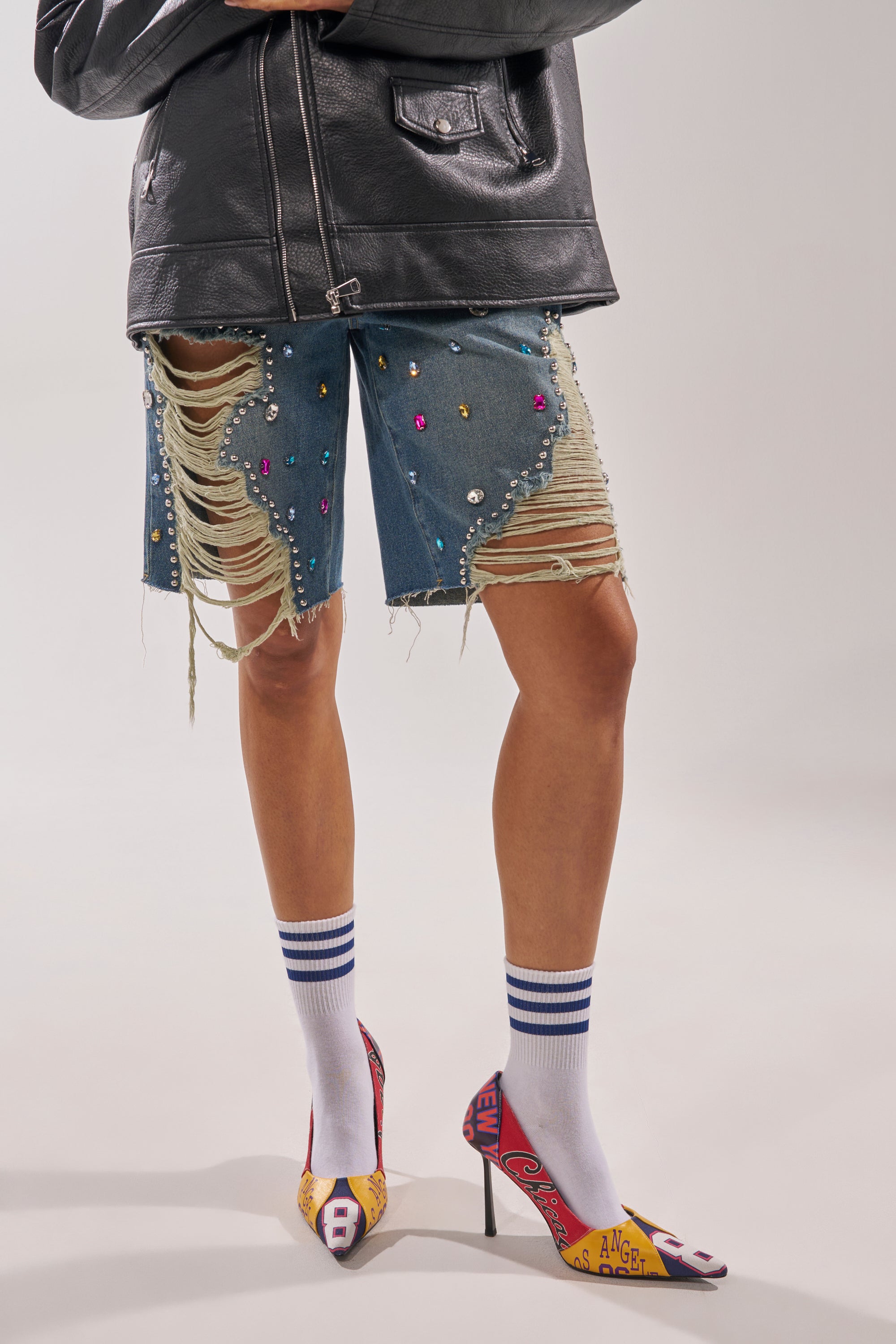 A person wearing a black leather jacket, ripped denim shorts adorned with colorful gemstones and pearls, white socks with blue stripes, and AZALEA WANG MULTI PRINTED STILETTO POINTED TOE PUMPS featuring bold patchwork graphics. 3