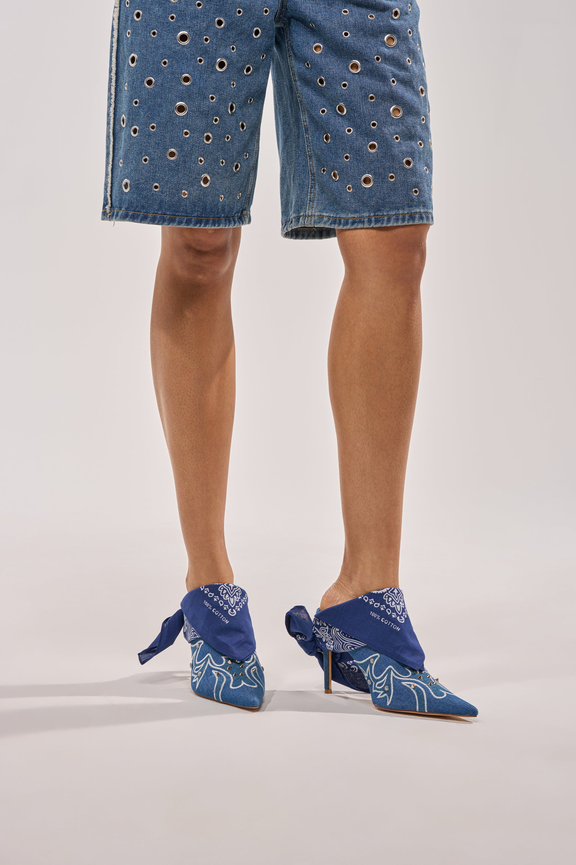 A person stands against a plain background wearing knee-length blue grommet-detailed denim shorts and the AZALEA WANG TAVINE DENIM MULE PUMP WITH BANDANA DETAIL. 1