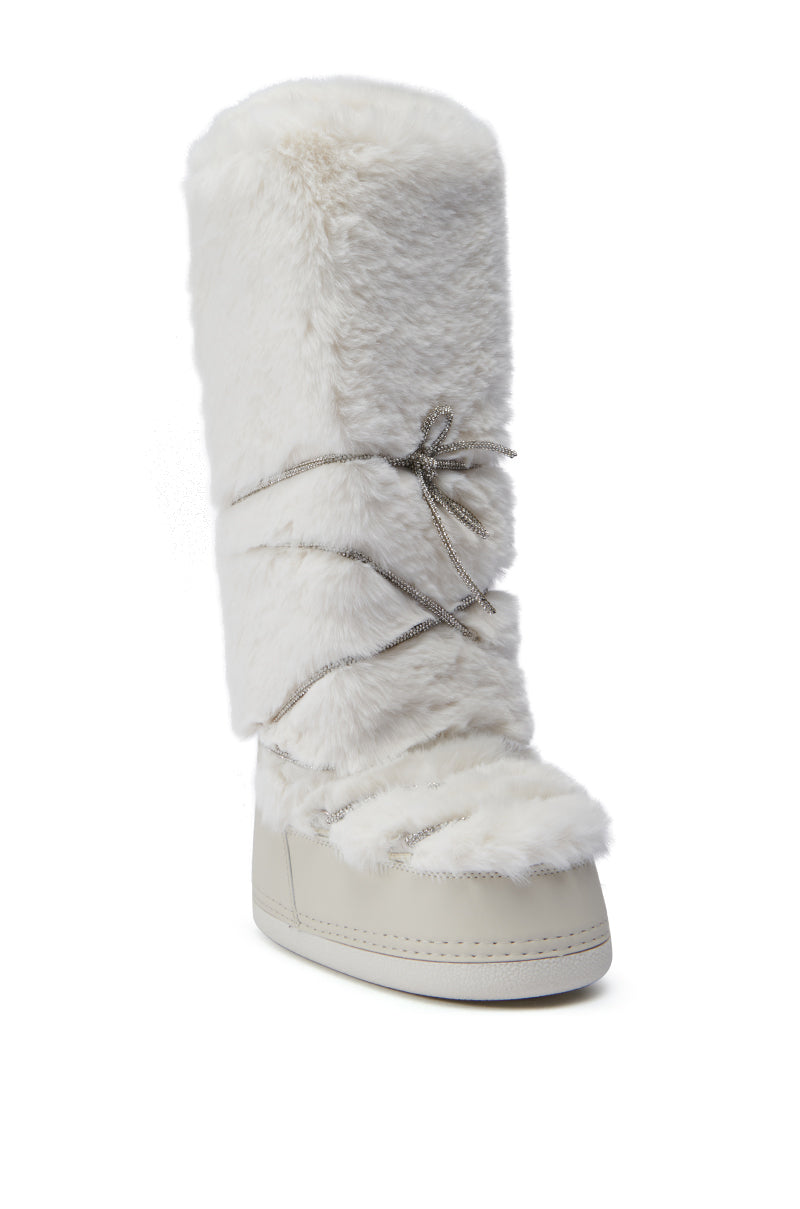 The AZALEA WANG TAYTON Beige Fuzzy Flat Knee High Boot features a soft faux fur exterior in white with a rounded toe. Decorative crisscrossing silver laces and rhinestone embellishments add sparkle to its design. The boot reaches up to the knee and is displayed against a plain white background with a flat sole for comfort.
