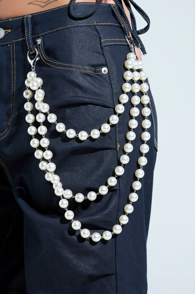 A person wears dark jeans with a stylish "STYLE ME UP PEARL PANTS CHAIN," featuring faux pearl strands and lobster claw closures. The large pearls drape elegantly, creating a striking contrast against the denim.