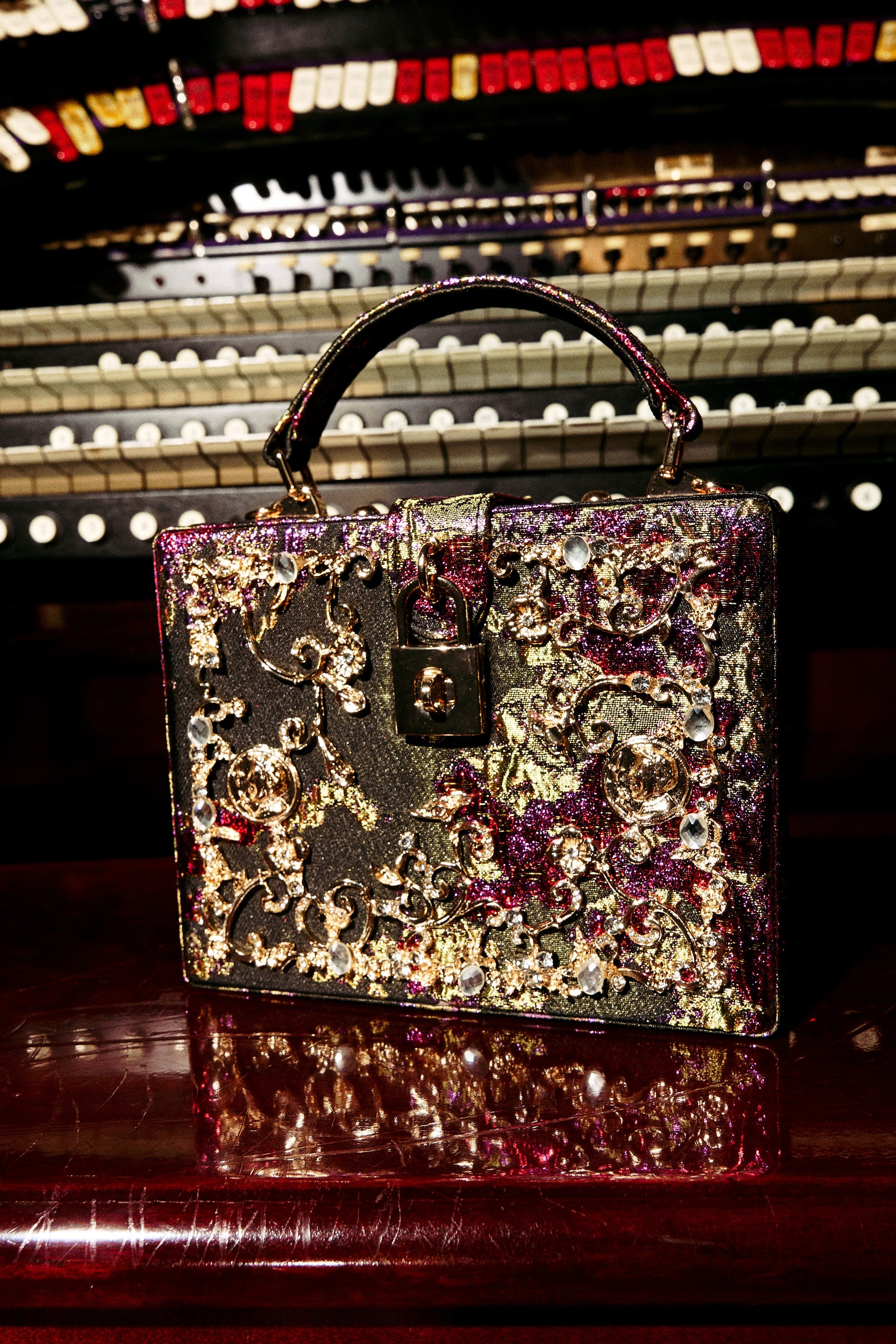 The BIG MONEY TRUNK BAG, a luxury handbag with gold, pink, and silver details, rests on a polished surface before an organ featuring multiple keyboards and vibrant buttons.