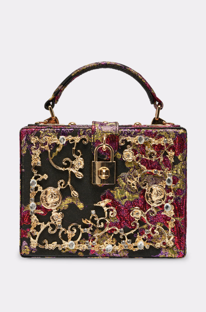 The BIG MONEY TRUNK BAG is a small rectangular bag with a top handle, featuring ornate gold embroidery, crystal embellishments, and a gold padlock on the front. Shown against black, red, green, and purple fabric backgrounds.