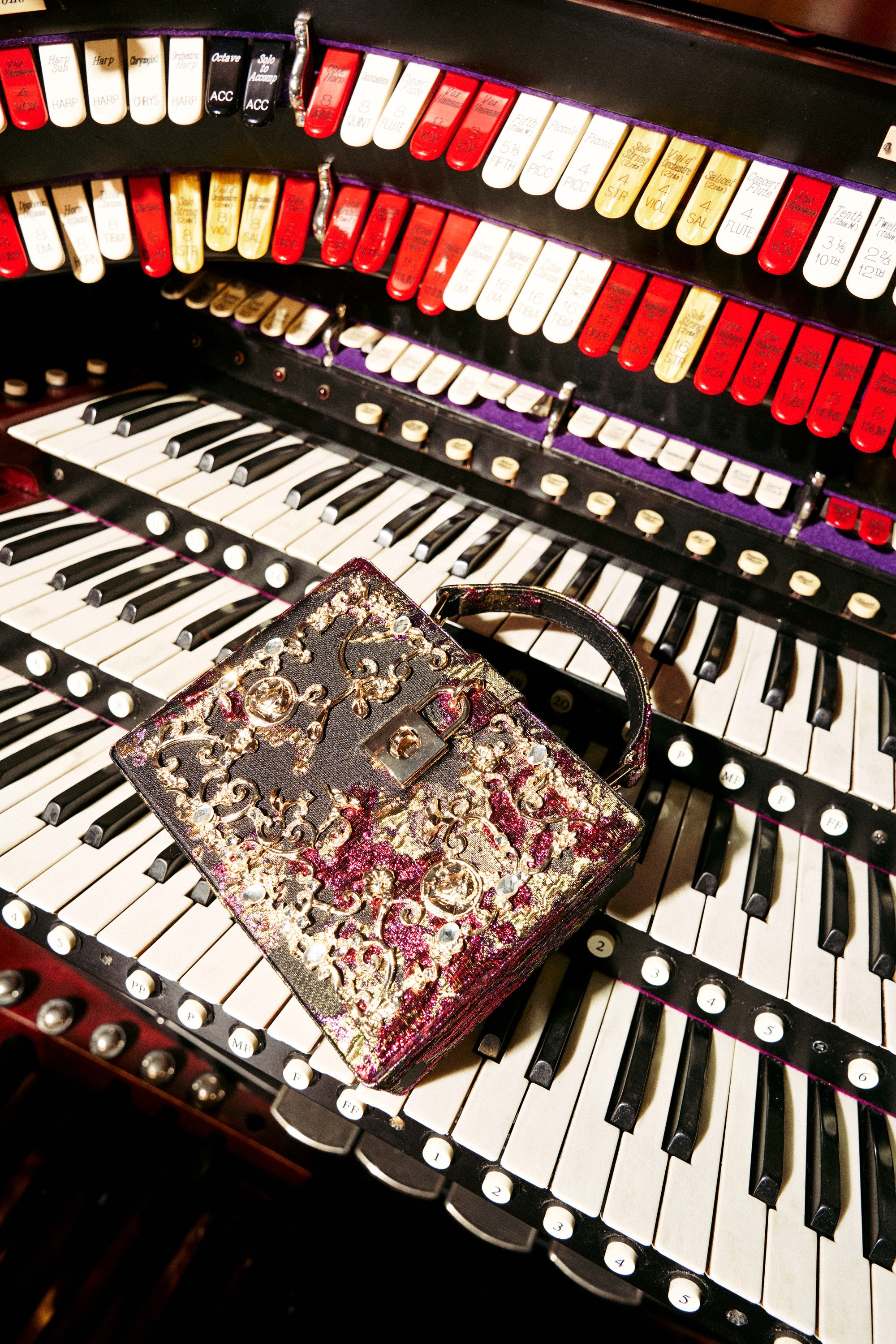 The BIG MONEY TRUNK BAG, adorned with gold and maroon embroidery, sits atop the keys of a grand organ surrounded by rows of vibrant buttons and switches.