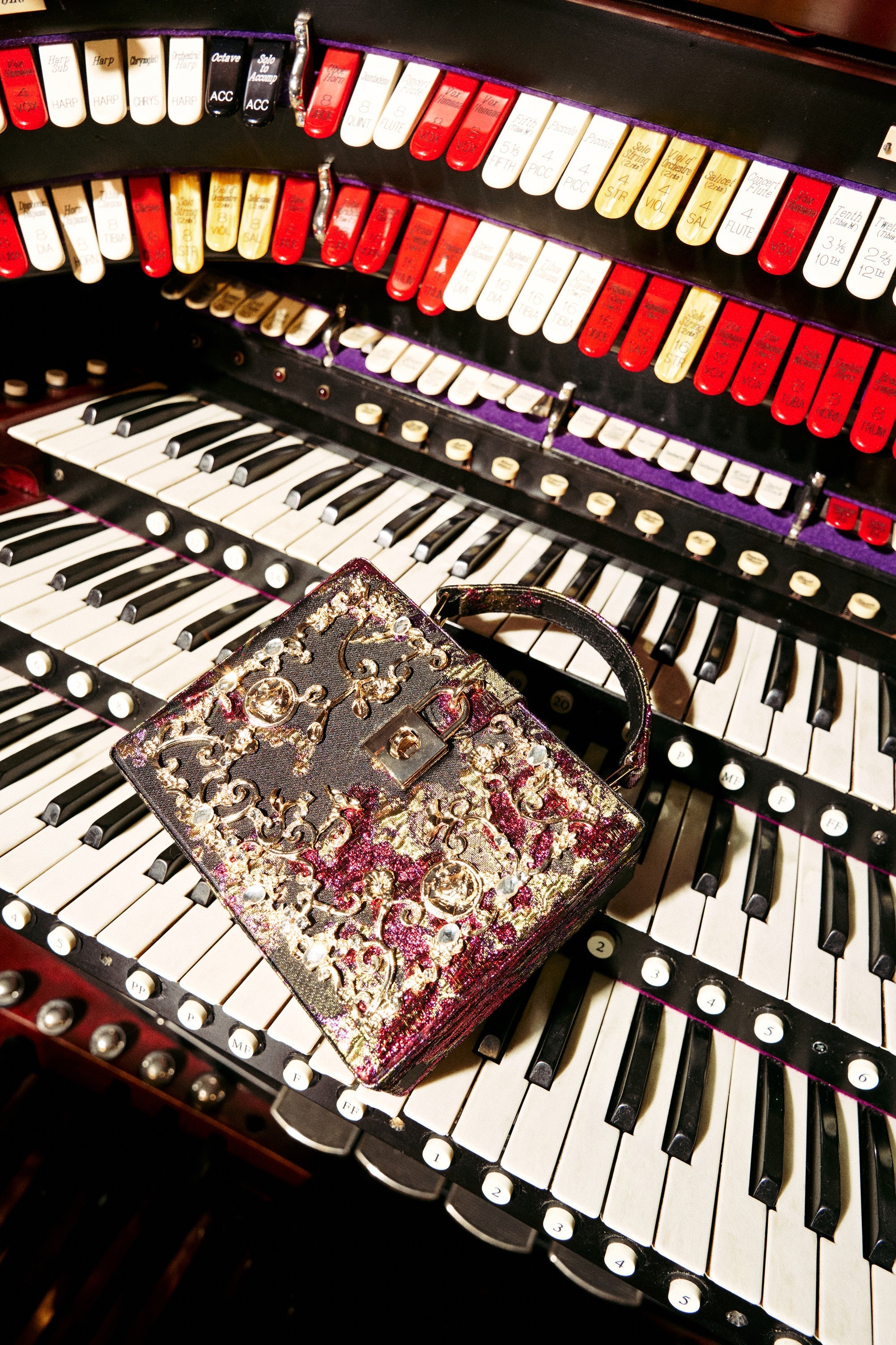 The BIG MONEY TRUNK BAG, adorned with gold and maroon embroidery, sits atop the keys of a grand organ surrounded by rows of vibrant buttons and switches. 5