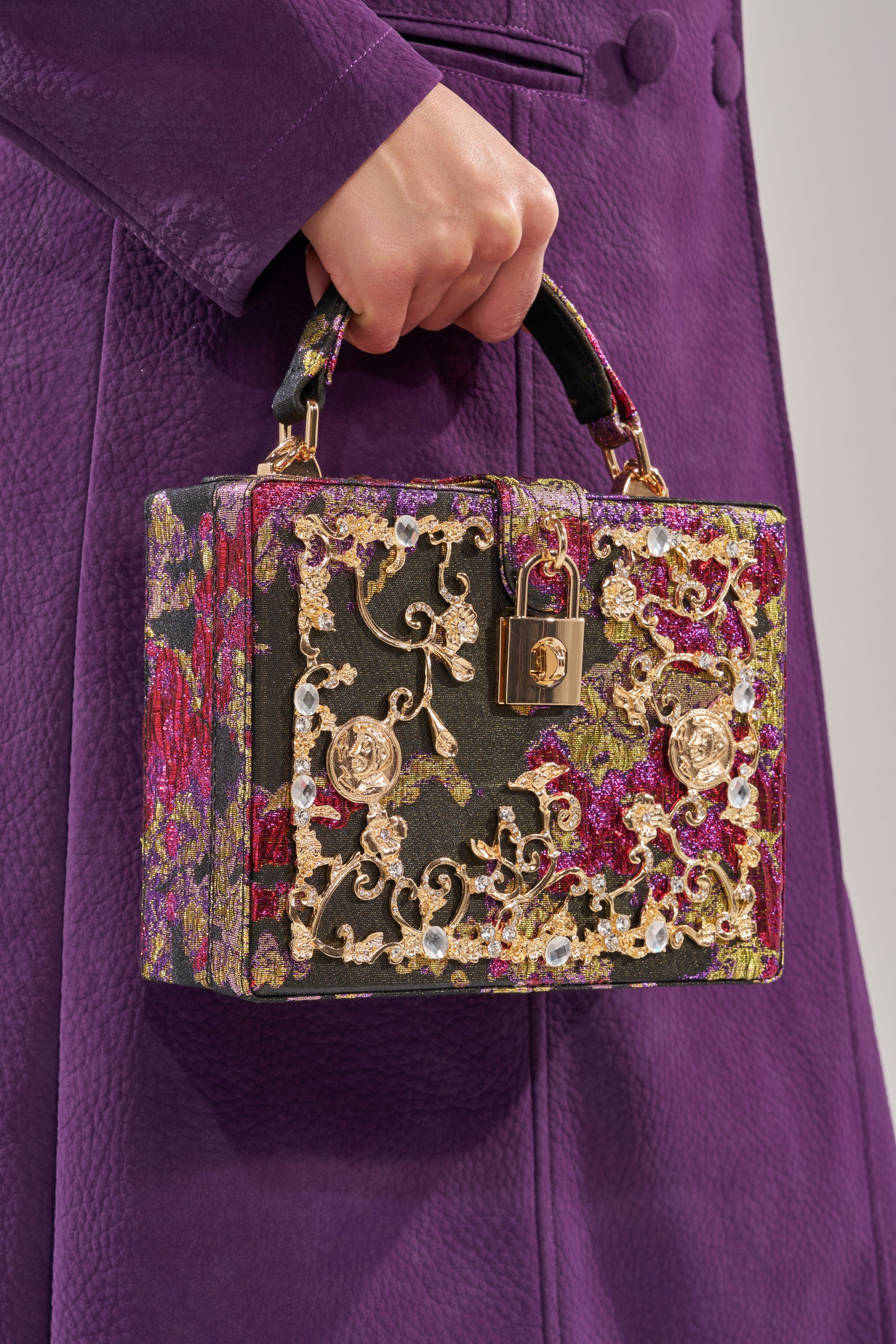 A person in a textured purple coat holds the BIG MONEY TRUNK BAG—a small luxury box bag with gold floral embroidery, gold clasp, and brocade designs in green, pink, and metallic tones.