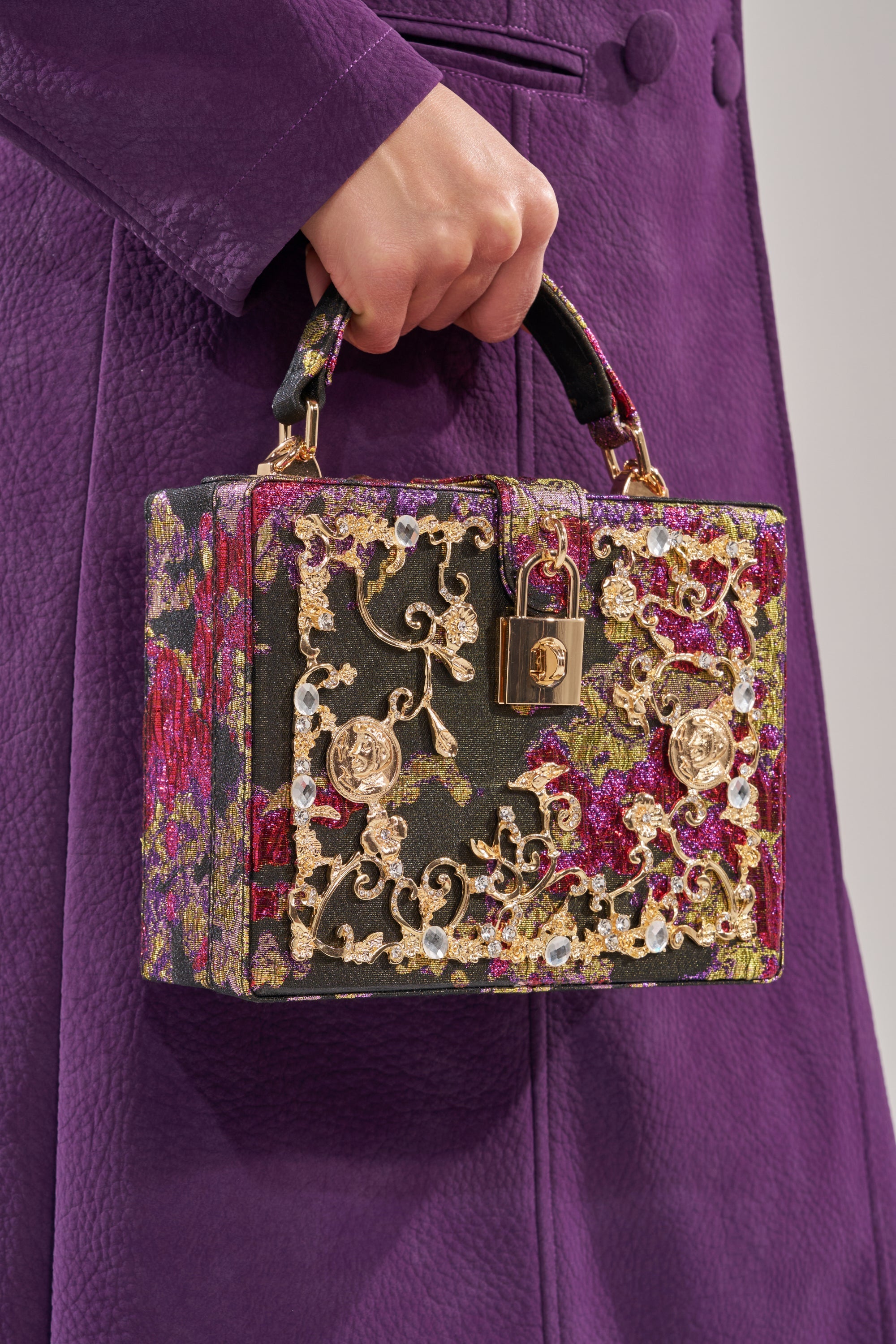 A person in a textured purple coat holds the BIG MONEY TRUNK BAG—a small luxury box bag with gold floral embroidery, gold clasp, and brocade designs in green, pink, and metallic tones. 1