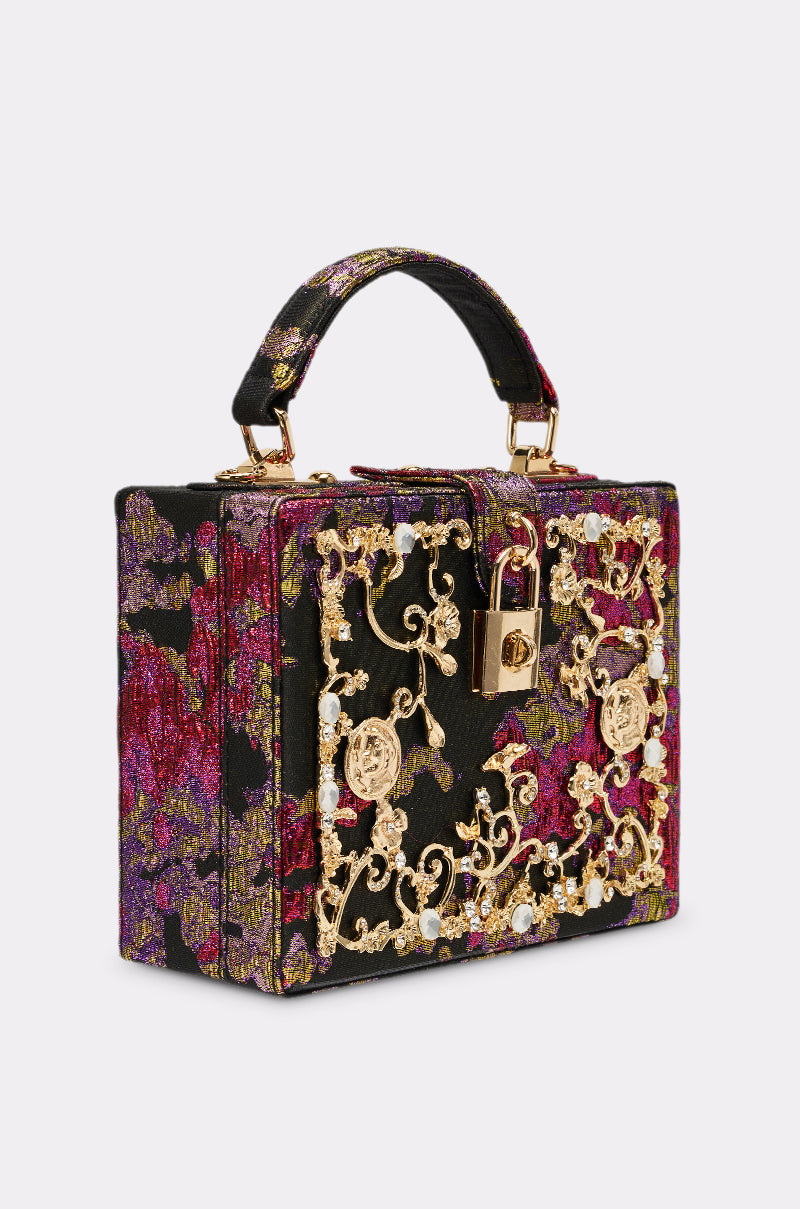 The BIG MONEY TRUNK BAG is a compact, box-shaped luxury bag crafted from red, purple, and gold floral fabric with ornate gold metal details, decorative gemstones, a front gold padlock, and a matching handle.