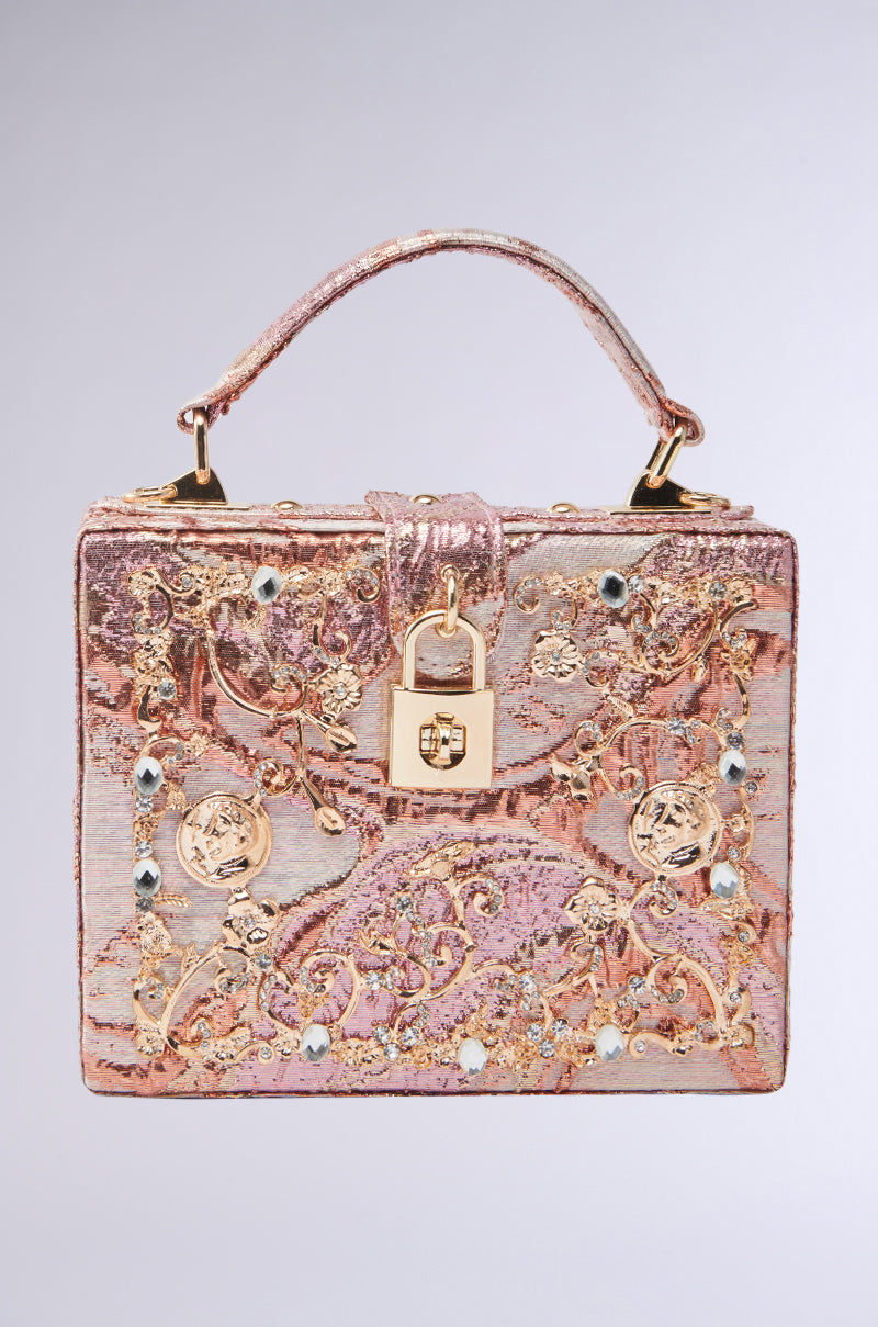 The BROCADE BABE TRUNK BAG is a luxurious rectangular bag with ornate pink and gold baroque brocade, metallic hardware, jewels, intricate pattern, top handle, gold clasp lock at the front, and a neutral light gray background.