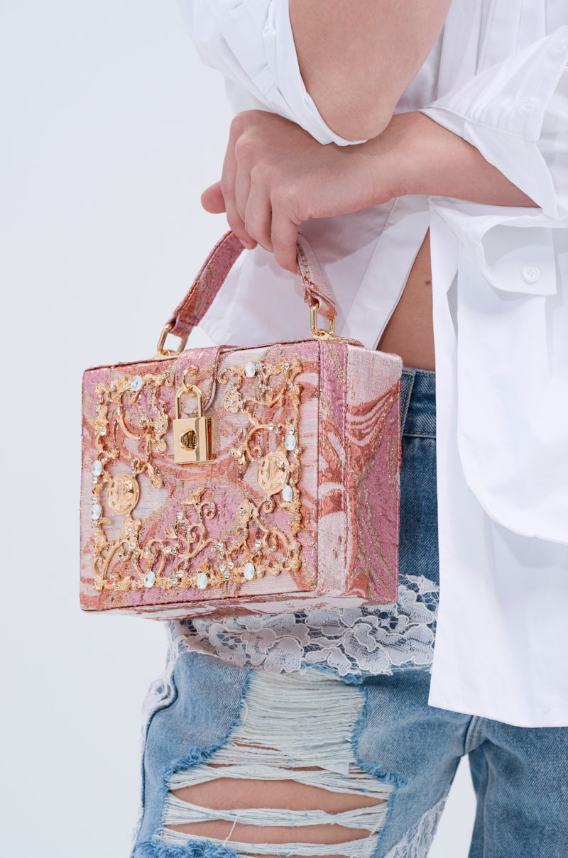 A person in a white shirt and ripped blue jeans with lace detail holds the BROCADE BABE TRUNK BAG, featuring ornate metallic brocade, faux gold hardware, and beaded embellishments.