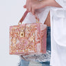 A person in a white shirt and ripped blue jeans with lace detail holds the BROCADE BABE TRUNK BAG, featuring ornate metallic brocade, faux gold hardware, and beaded embellishments.