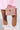 A person carries the BROCADE BABE TRUNK BAG, hugged in ornate gold decorations and metallic hardware. They're dressed in a pink textured ensemble, complemented by ivory ruffled socks and shiny pink shoes, against a plain white background.