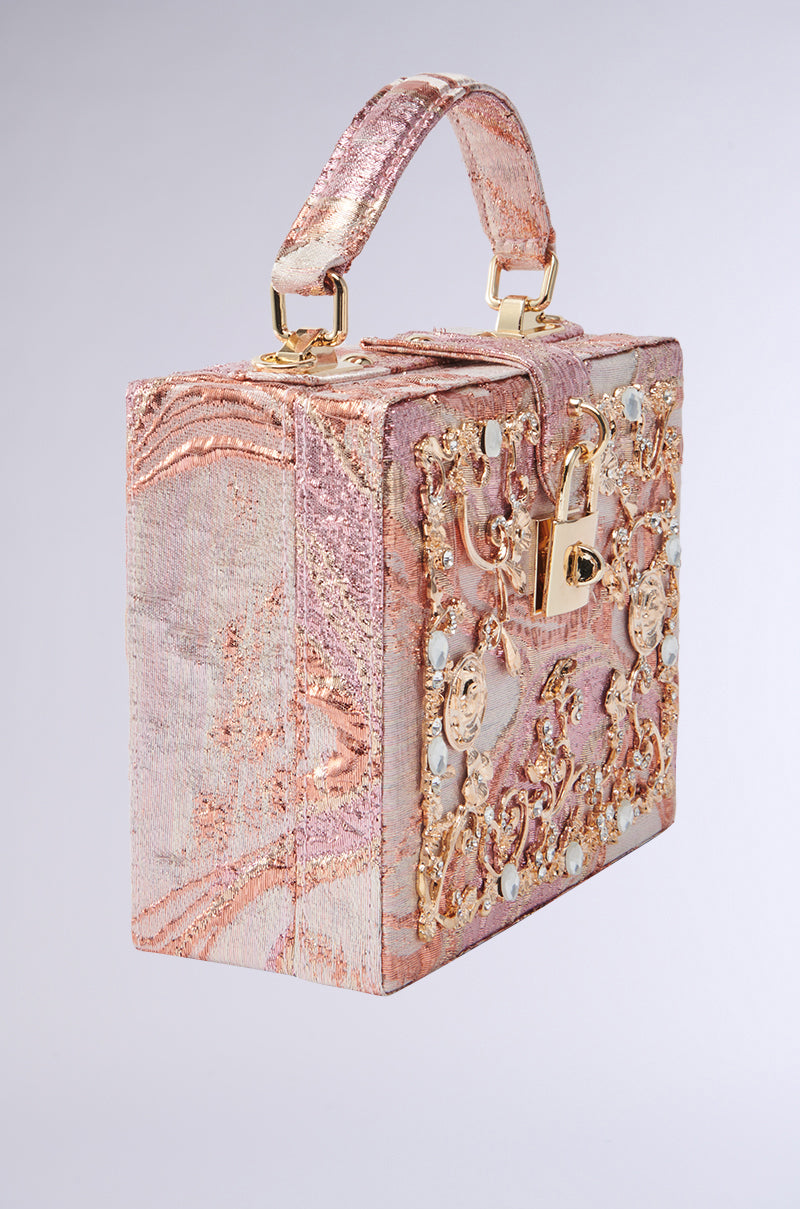 The BROCADE BABE TRUNK BAG is a square, ornate masterpiece with a structured handle and lock. It features a luxurious pink and gold texture, adorned with intricate patterns and embellishments, while metallic hardware elevates its sophisticated elegance.