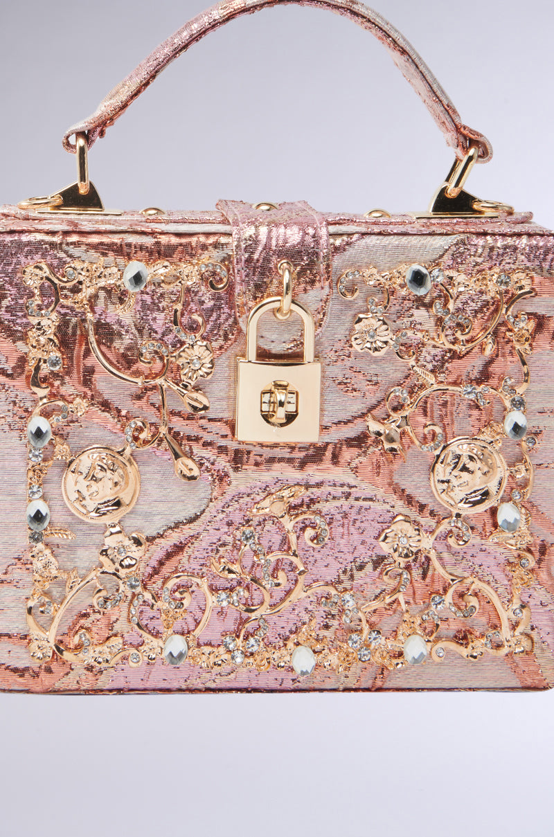 The BROCADE BABE TRUNK BAG is a luxurious trunk bag featuring intricate gold embroidery and gemstones, metallic hardware with a central decorative lock, a shimmering pink and gold texture, and a sturdy top handle.