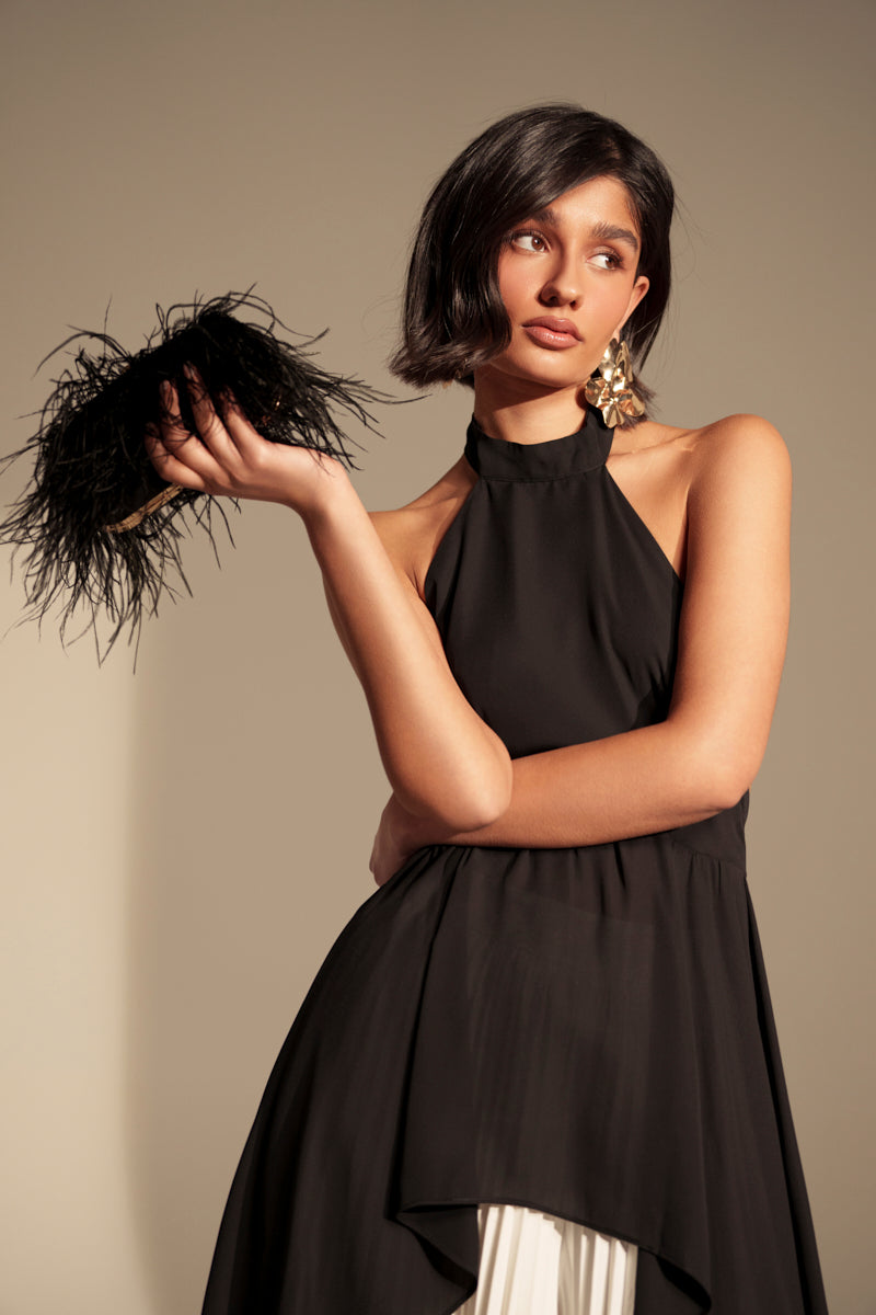 A woman holds the MIDNIGHT IN PARIS FEATHER CLUTCH in one hand, pairing it with a sleeveless black halter dress and large faux gold earrings, looking to the side against a plain background.