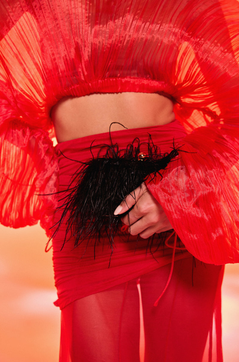 Wearing a sheer, pleated red top and skirt, this person holds the MIDNIGHT IN PARIS FEATHER CLUTCH against a warm, orange-toned background. 0