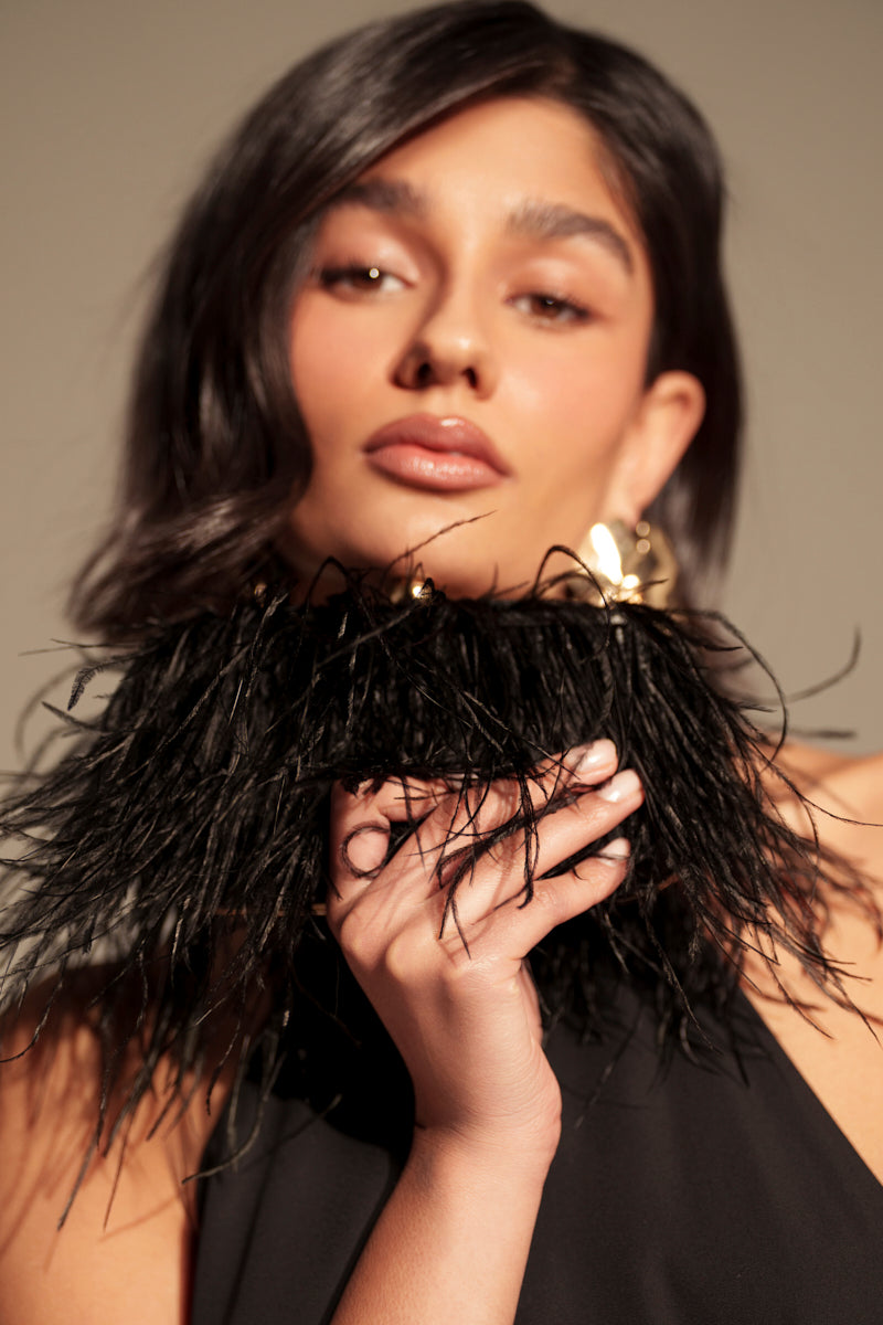 A woman with wavy dark hair poses in a halter neck black outfit with a dramatic feather collar, holding the MIDNIGHT IN PARIS FEATHER CLUTCH featuring faux gold metal details near her face against a neutral background. 2
