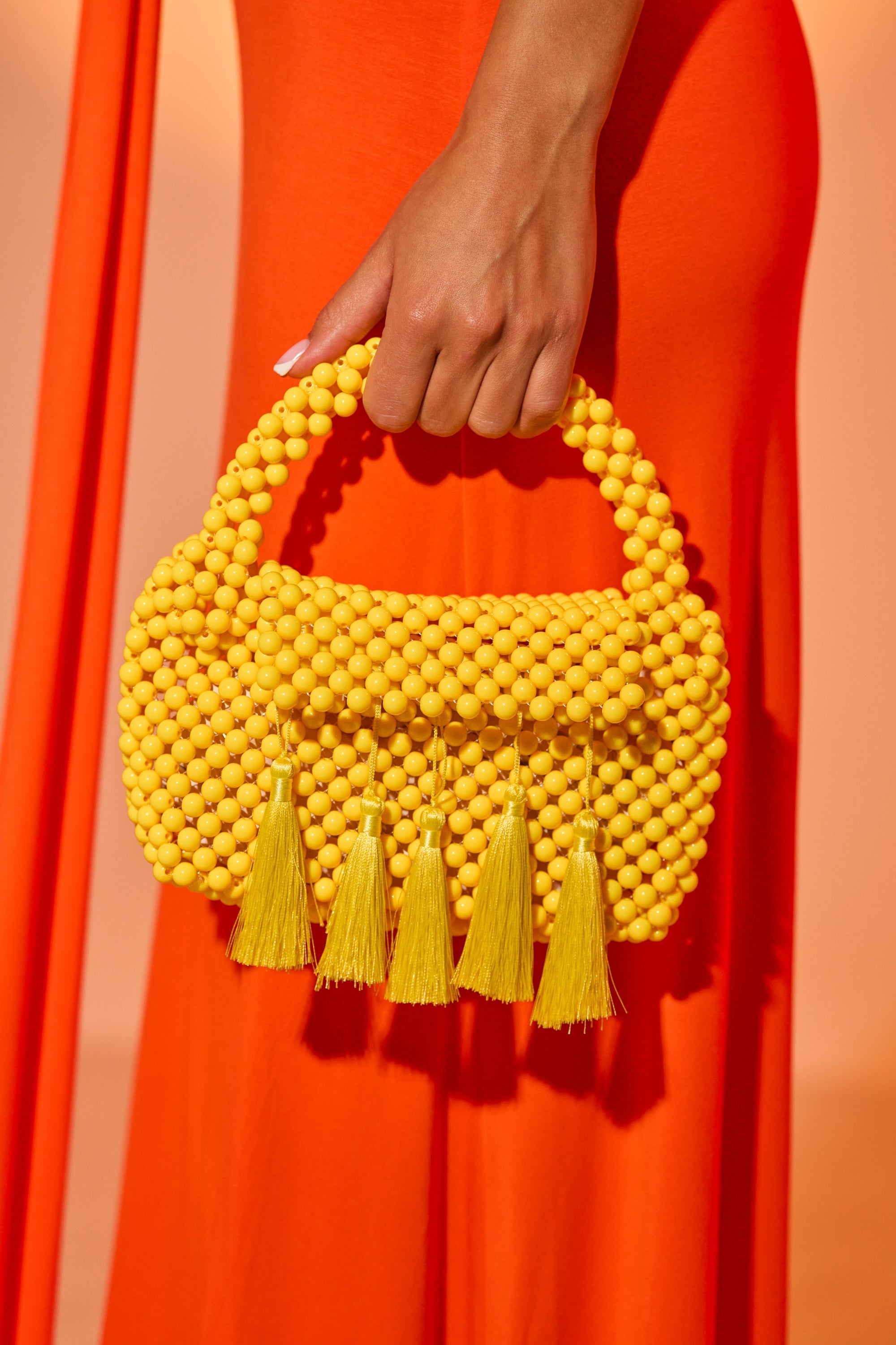A person in a bright orange dress holds the IN THE SUN BEADED BAG—a yellow resin beaded handbag with a rounded handle and playful tassels, perfect as a vibrant summer accessory. 0