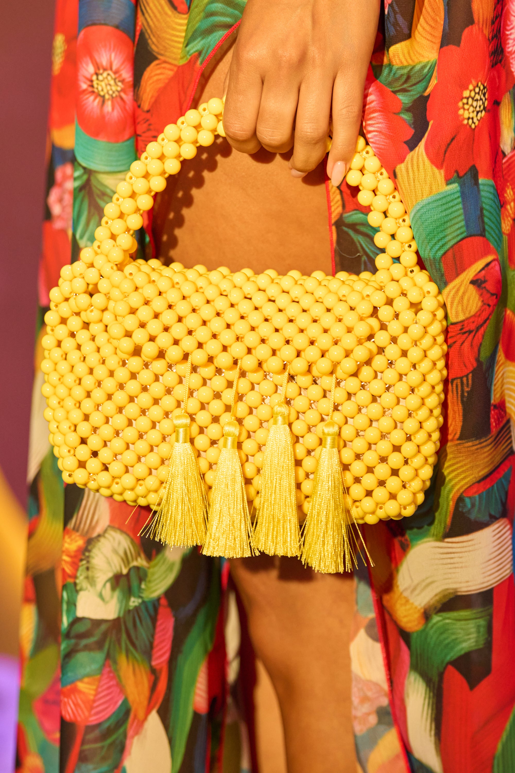 A hand holds the IN THE SUN BEADED BAG—a yellow handbag with a beaded handle and four tassels—adding a charming, eye-catching touch to any summer outfit against a colorful floral dress. 0