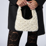 A person wearing a pinstriped blazer over a crocheted top and black lace flared pants is holding an OLD MONEY PEARL EMBELLISHED PURSE. The small, white satin bag features faux pearl beading and a braided handle, while the person's face remains out of sight.