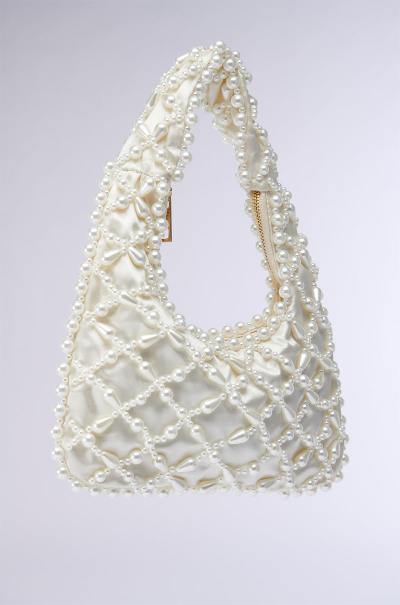 Introducing the OLD MONEY PEARL EMBELLISHED PURSE, a stylish accessory adorned with faux pearl beading in a crisscross pattern. This elegant white purse features a single handle with intricate pearl detailing, has a slight sheen, and includes a zipper closure at the top. The background is plain and light-colored, perfectly highlighting its sophisticated design.