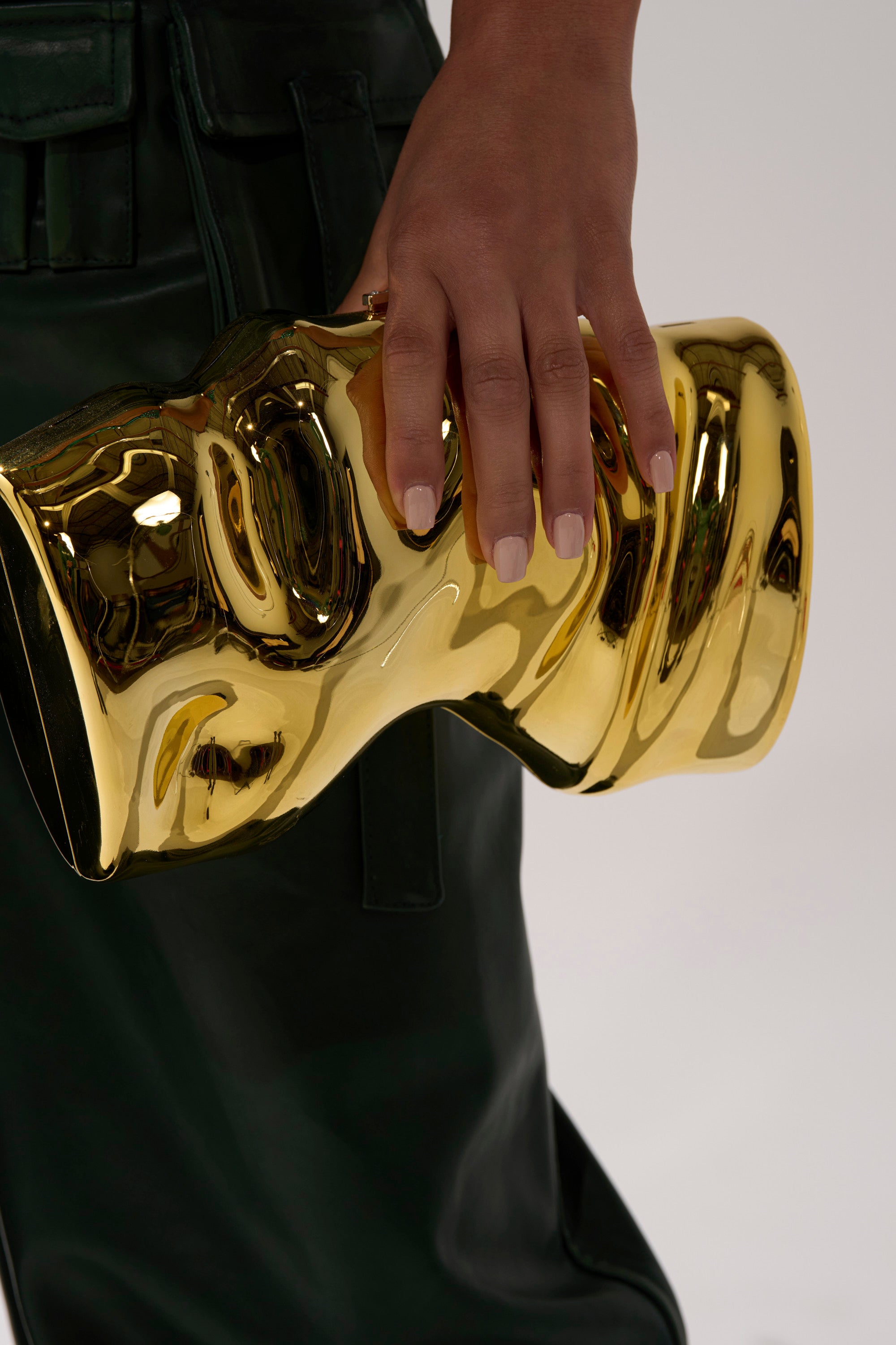 A person with manicured nails holds the FOR THE TIMES CLUTCH—a shiny, gold evening bag with a sculptural design and rhinestone clasp—while wearing dark green pants.