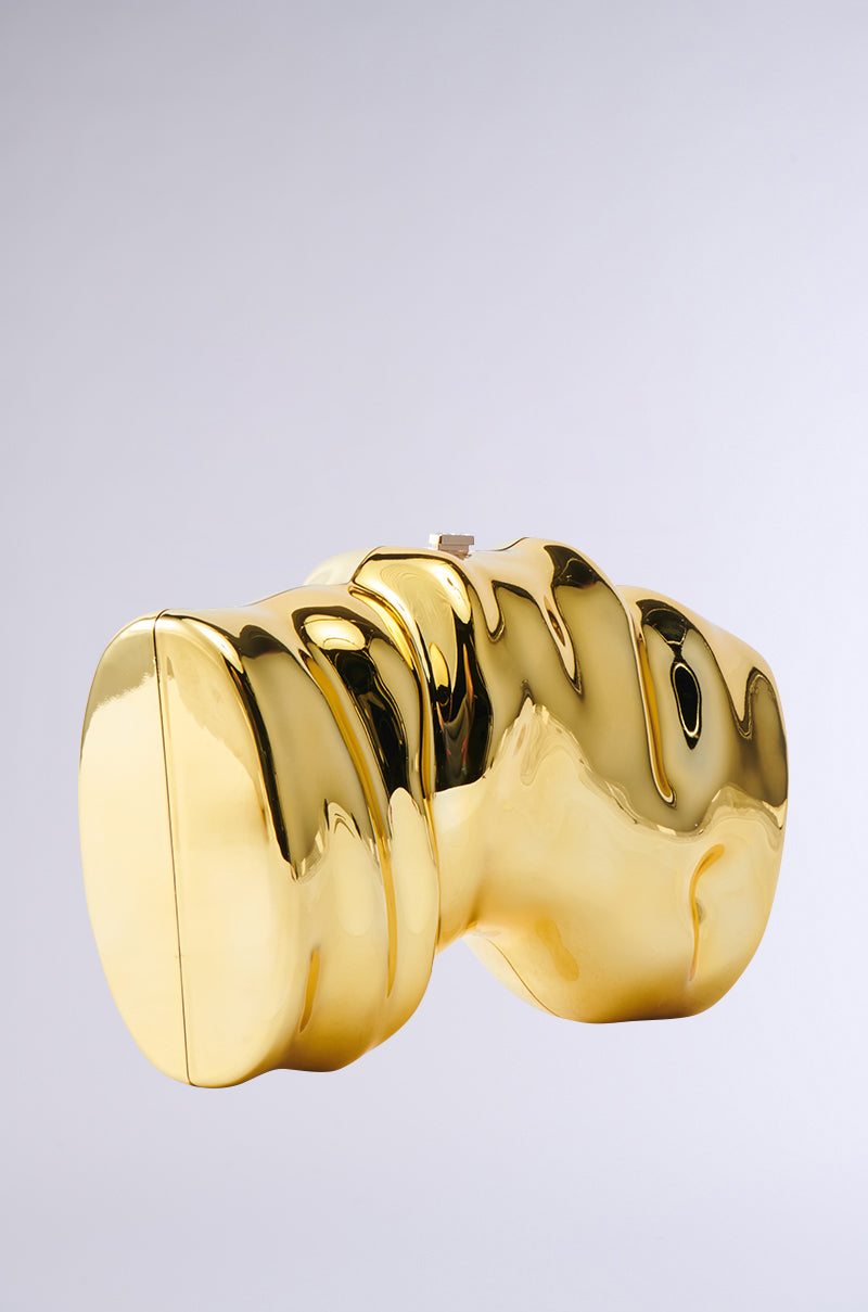 The FOR THE TIMES CLUTCH is a shiny gold, abstract-shaped accessory with smooth, wavy contours, making a sculptural statement as it’s elegantly displayed against a plain light gray background.
