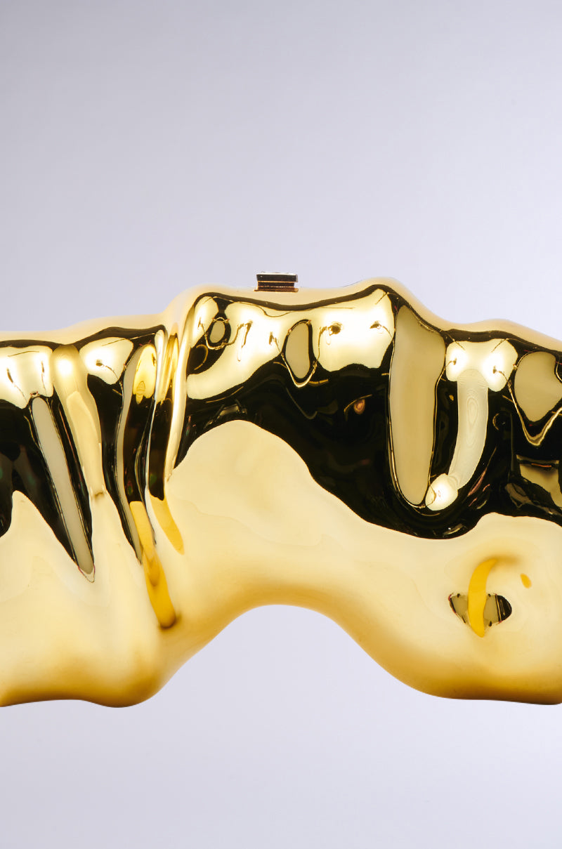A close-up of a shiny, abstract gold sculpture with smooth curves evokes the look of a sculptural statement piece, reminiscent of the FOR THE TIMES CLUTCH, set against a plain light gray background.