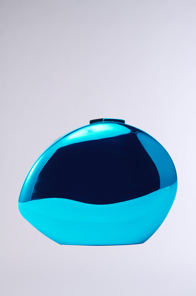 The NO BLUES CHROME CLUTCH, an oval-shaped aquatic blue metallic object, has a smooth surface that elegantly reflects light while resting on a plain, light gray background, exuding a serene yet captivating presence.