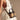A woman in a white, halter-neck, eyelet dress carries the TAKE ME OUT WICKER PURSE—a small black and tan wicker bag with a shoulder strap and gold-tone hardware. Soft lighting enhances the dress's texture and vintage charm.