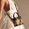 A woman in a white, halter-neck, eyelet dress carries the TAKE ME OUT WICKER PURSE—a small black and tan wicker bag with a shoulder strap and gold-tone hardware. Soft lighting enhances the dress's texture and vintage charm.