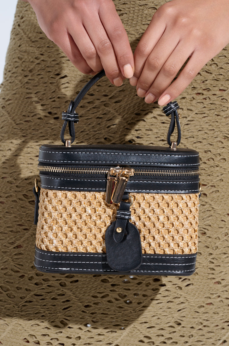Close-up of hands holding the TAKE ME OUT WICKER PURSE, featuring black faux leather trim and a gold zipper. The person wears a textured, olive-green garment.