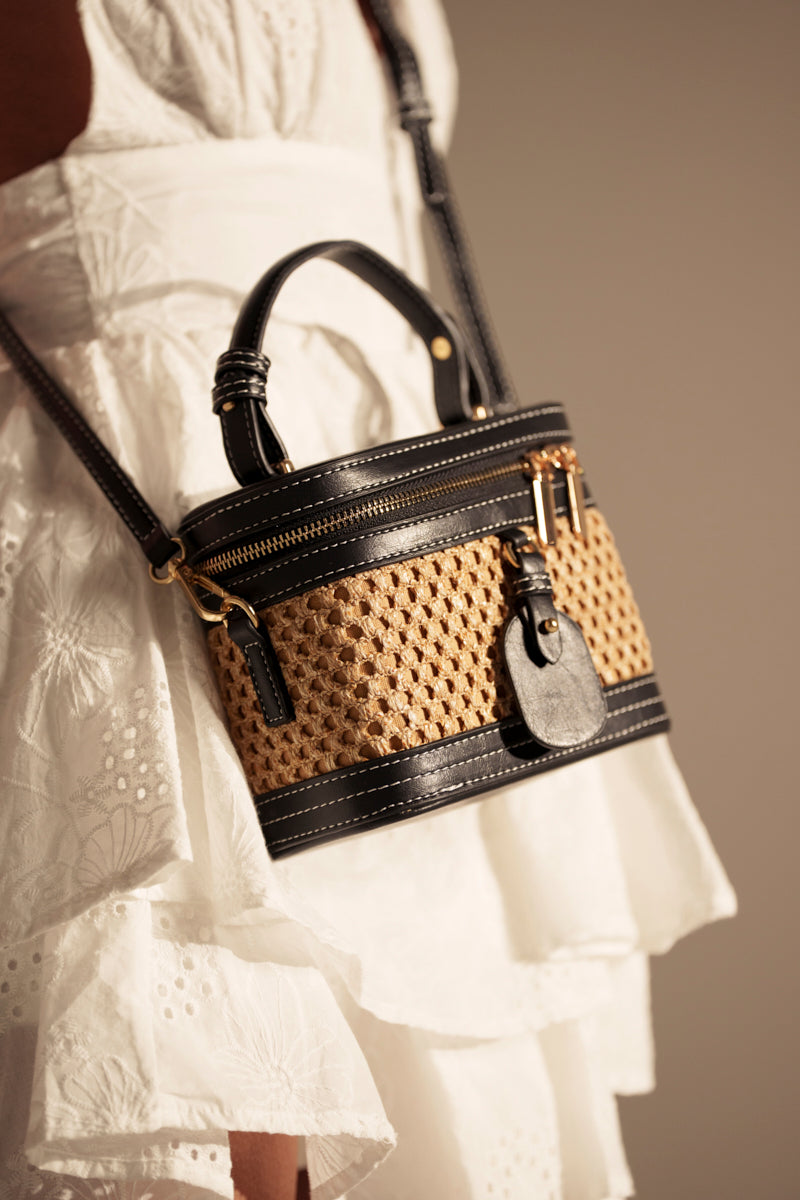 A person in a white eyelet dress carries the TAKE ME OUT WICKER PURSE, a small rattan bag with black faux leather trim, gold hardware, a top handle, and worn crossbody.