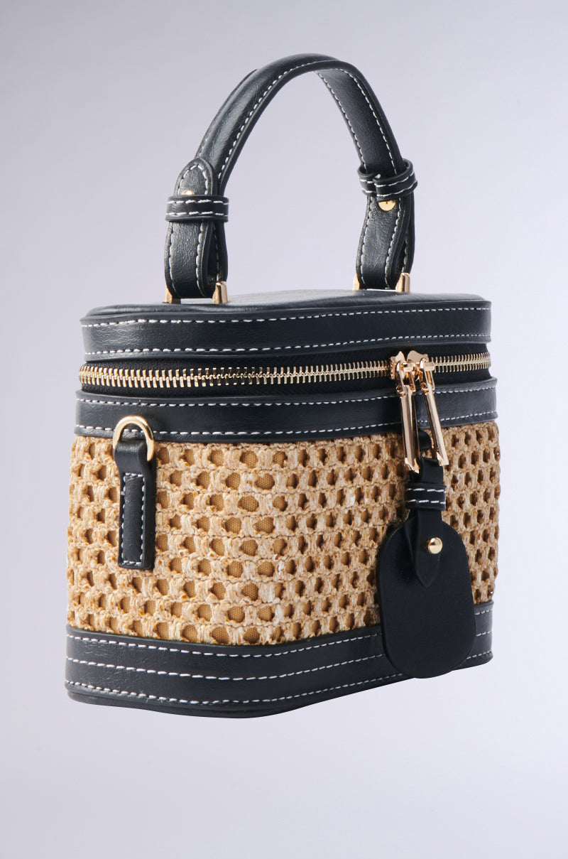 The "TAKE ME OUT WICKER PURSE" combines a stylish rattan design with faux leather trim, showcasing a woven texture accented by black leather details. Its rounded rectangular shape features a zipper closure, gold hardware, and a sleek black handle, all set against a plain gray background for timeless sophistication.