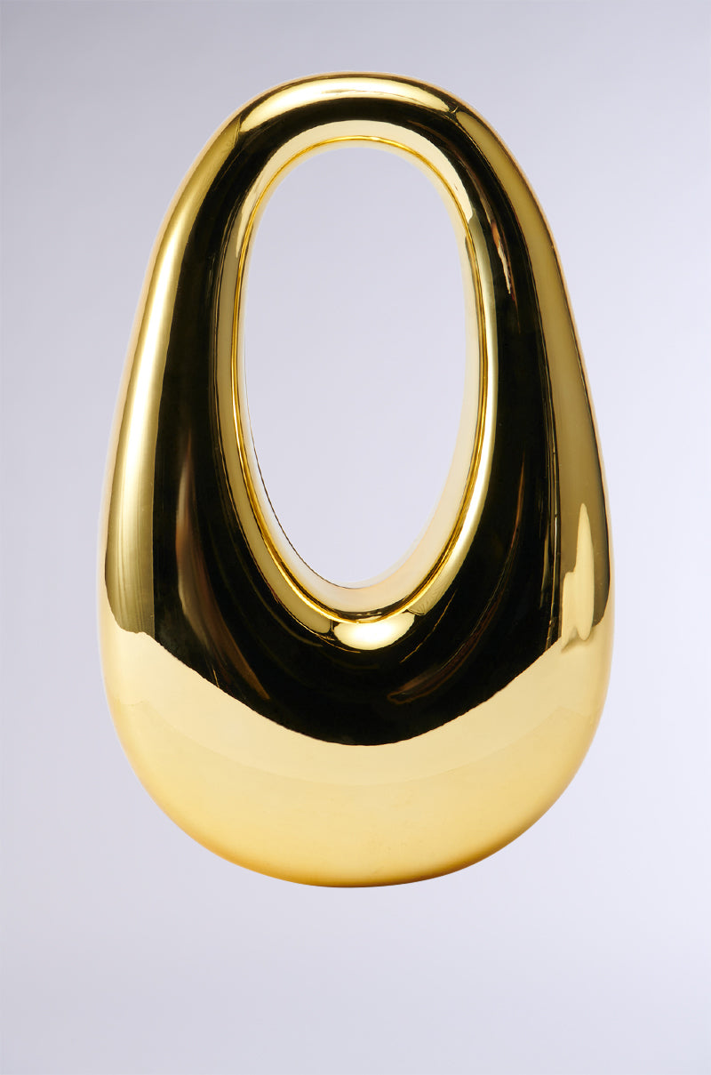 A polished, golden sculpture featuring an egg-shaped design with an oval cut-out at its center evokes the chic elegance of a hardshell frame bag. Positioned against a simple gray background, its highly reflective and glossy surface captures the allure of the GOLDEN HOUR CHROME PURSE.