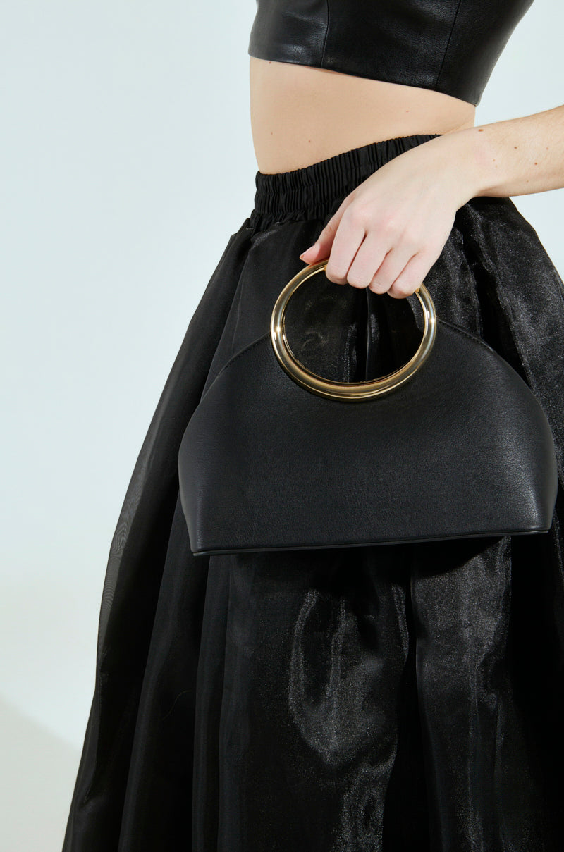 The torso of a person in a black leather crop top and satin skirt is seen against a plain background, holding the I AM LUXE PURSE with large gold circular handles, capturing quiet luxury. 1