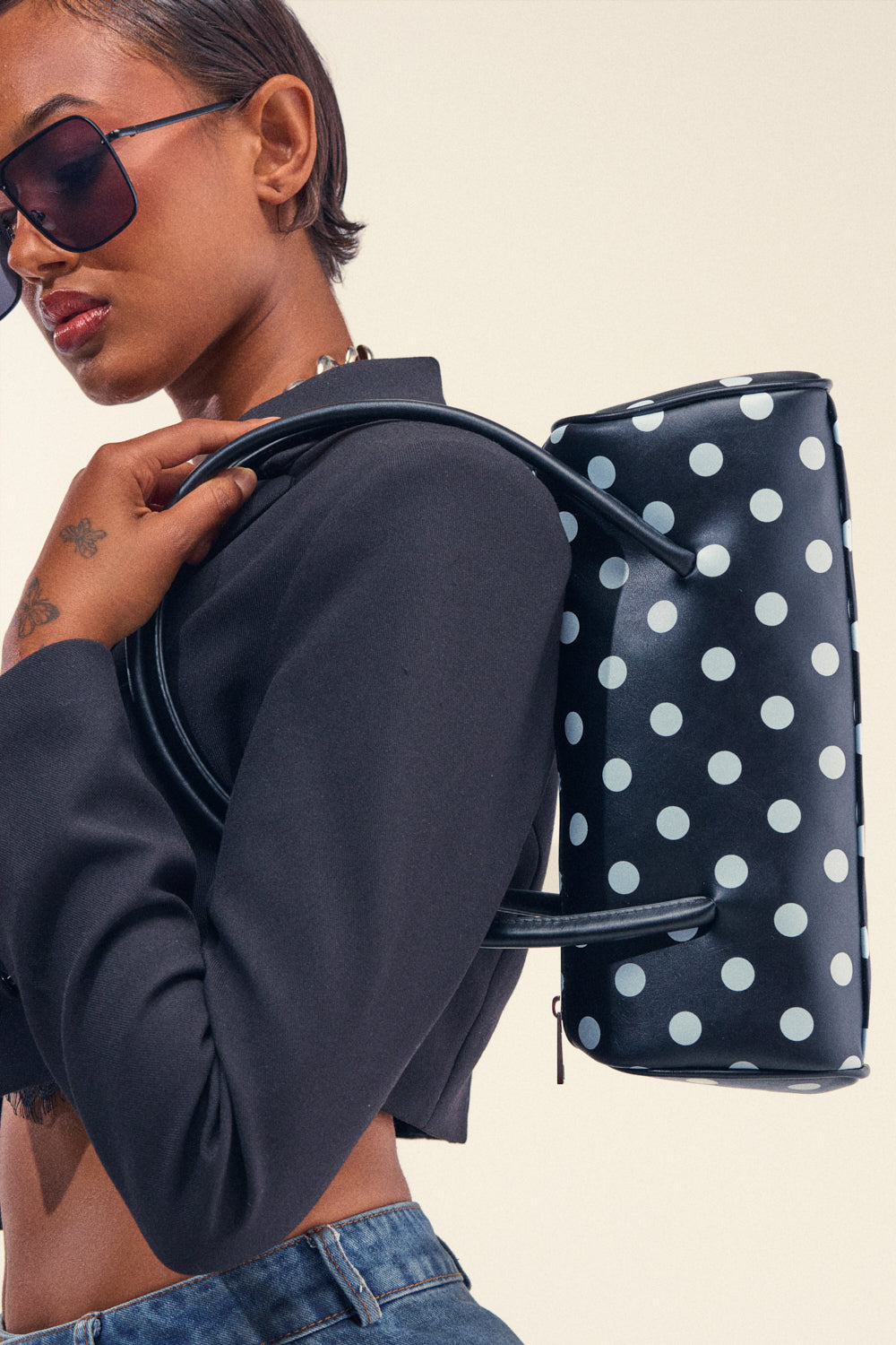 A person in sunglasses and a black cropped jacket poses with the SPOT IT TREND PURSE, a chic faux leather bag featuring a playful polka dot print, held over their shoulder against a plain background.