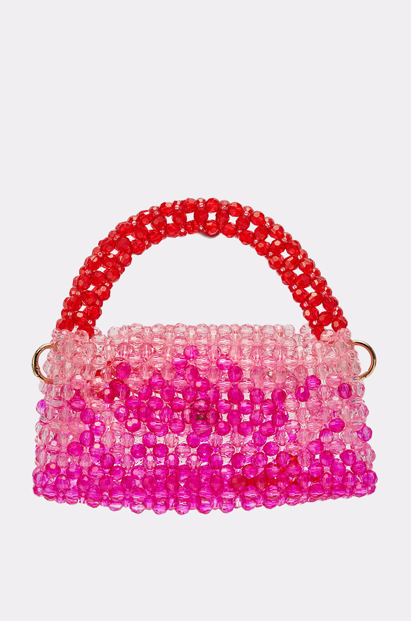 The IN THE TROPICS BEADED BAG is a small bag crafted from translucent pink and magenta beads, featuring an ombré red and pink beaded handle, shown on a plain white background. 1