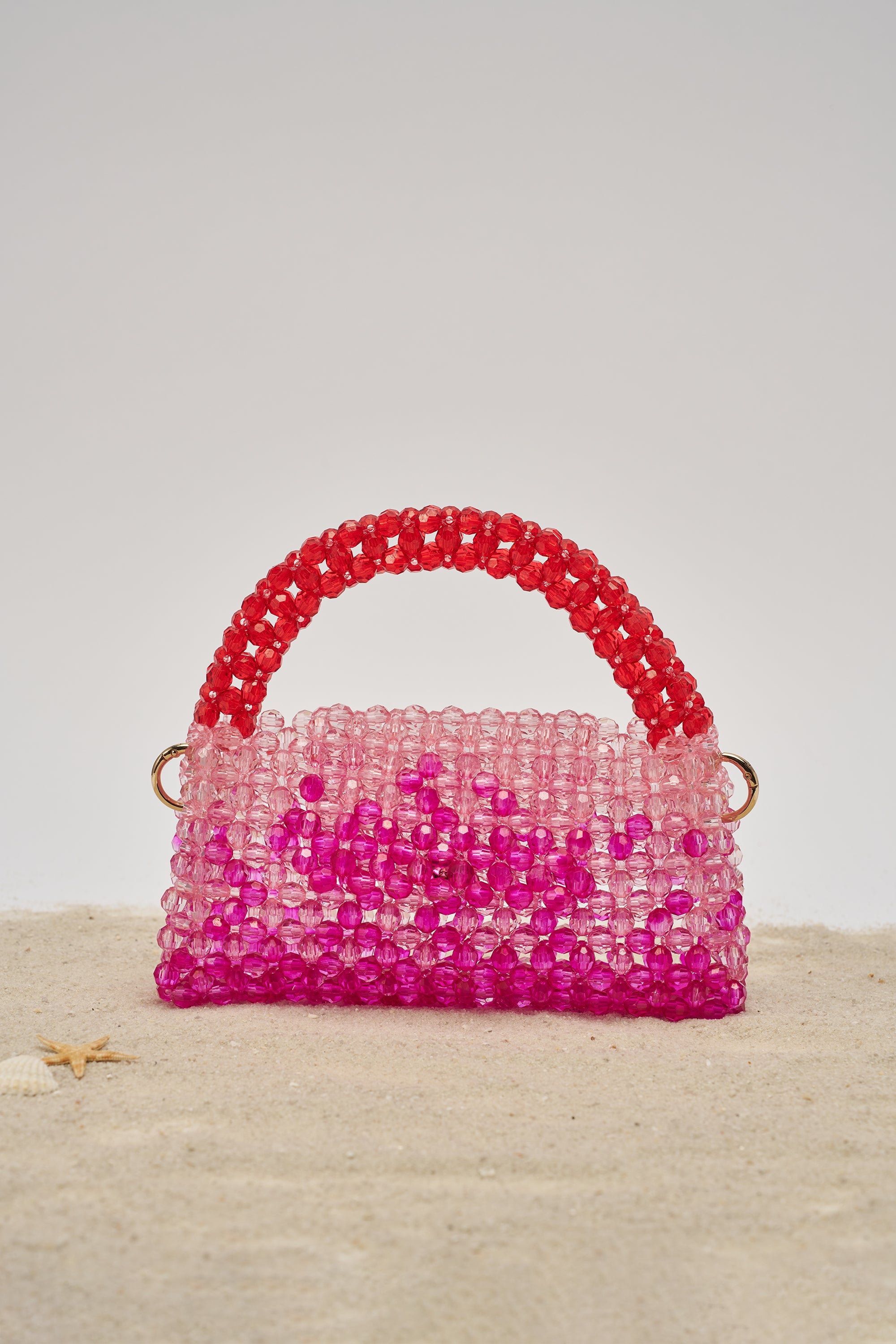 The IN THE TROPICS BEADED BAG, featuring a body that fades from light pink to vibrant magenta and an ombré red and pink beaded handle, rests on sand beside a tiny starfish and shell. 0