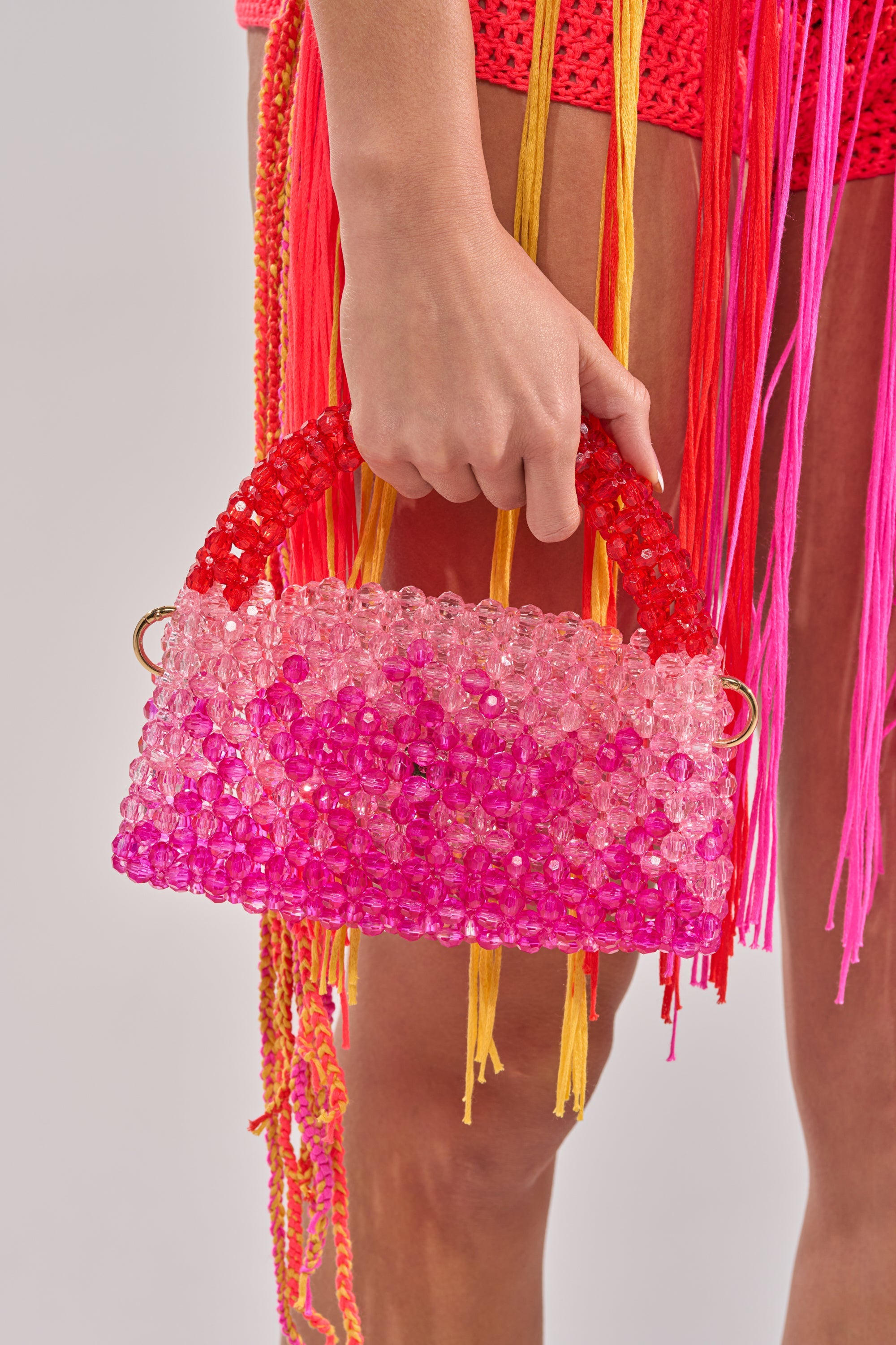A person holds the IN THE TROPICS BEADED BAG in ombré red-pink, paired with a crocheted outfit with pink and yellow fringe. The simple, light background draws attention to the bag’s vibrant colors and textures. 2