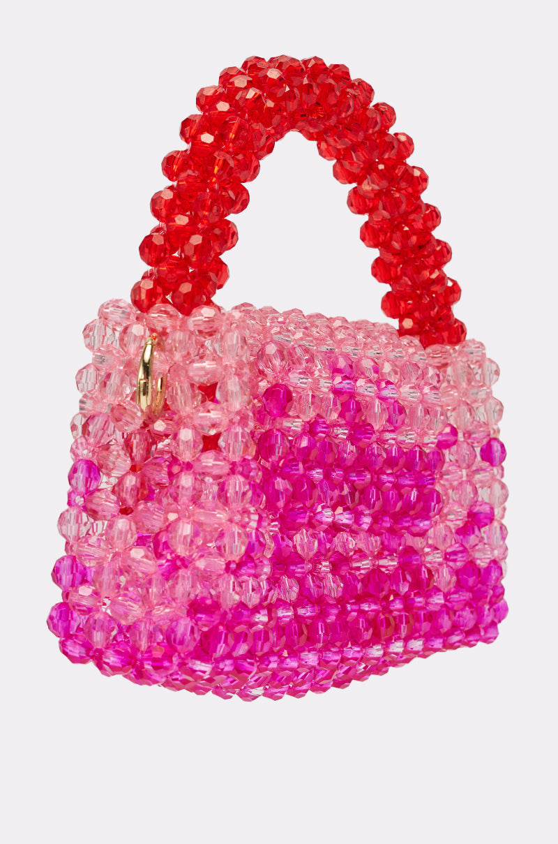 The IN THE TROPICS BEADED BAG is a small, ombré red and pink beaded bag with a chunky red handle and gold clasp, crafted from translucent pink and magenta beads, shown against a plain white background. 3