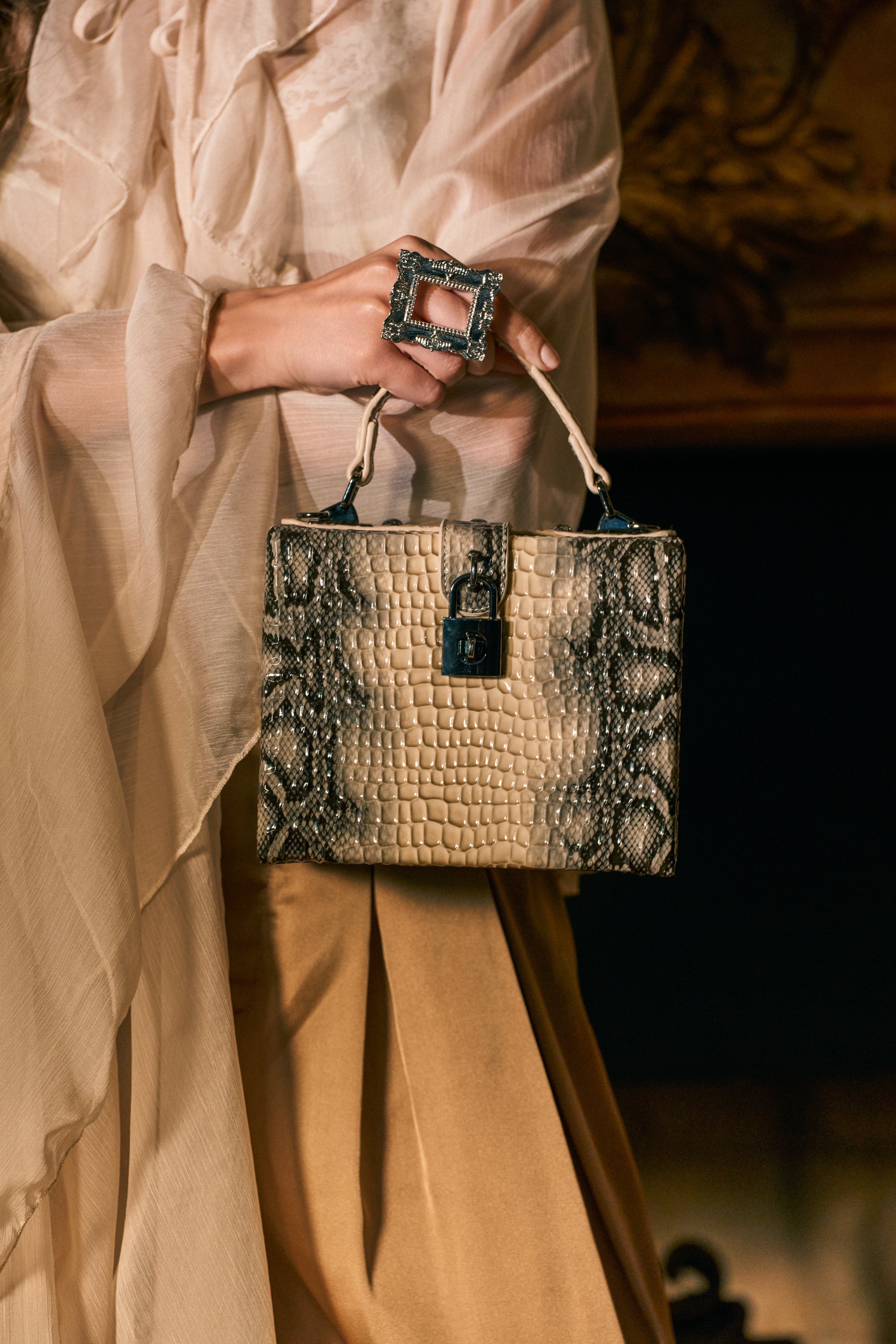 A person in a sheer, cream blouse holds the LITTLE SNAKE HANDBAG—a chic mini box bag featuring a bold snake print pattern and structured top handle. They wear a large, square statement ring on one hand.