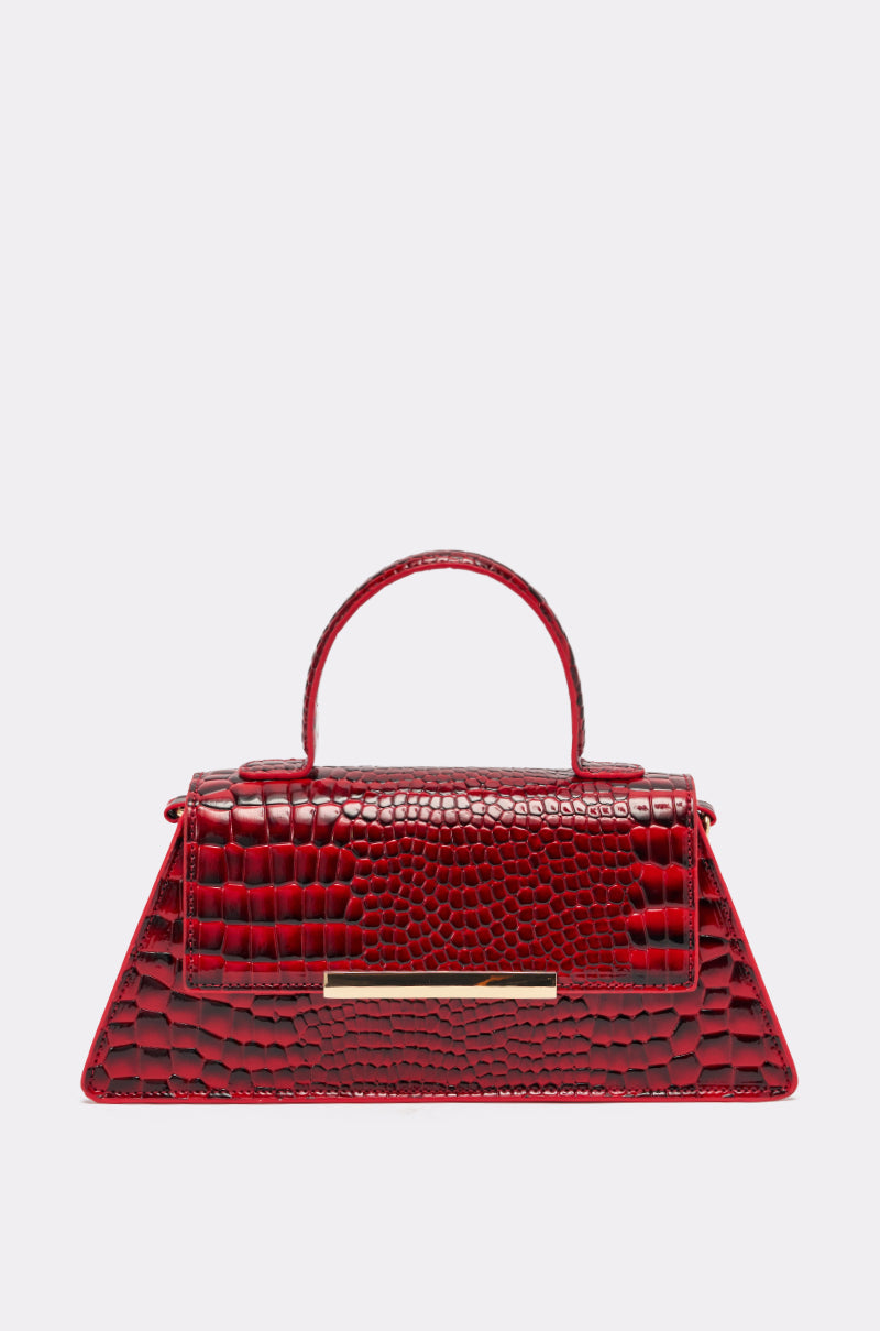 The DAY FAUX CROC PURSE is a cherry red, trapezoid-shaped handbag with crocodile-textured vegan leather, a top handle, and a gold horizontal bar accent on the front flap, shown against a plain light background. 0