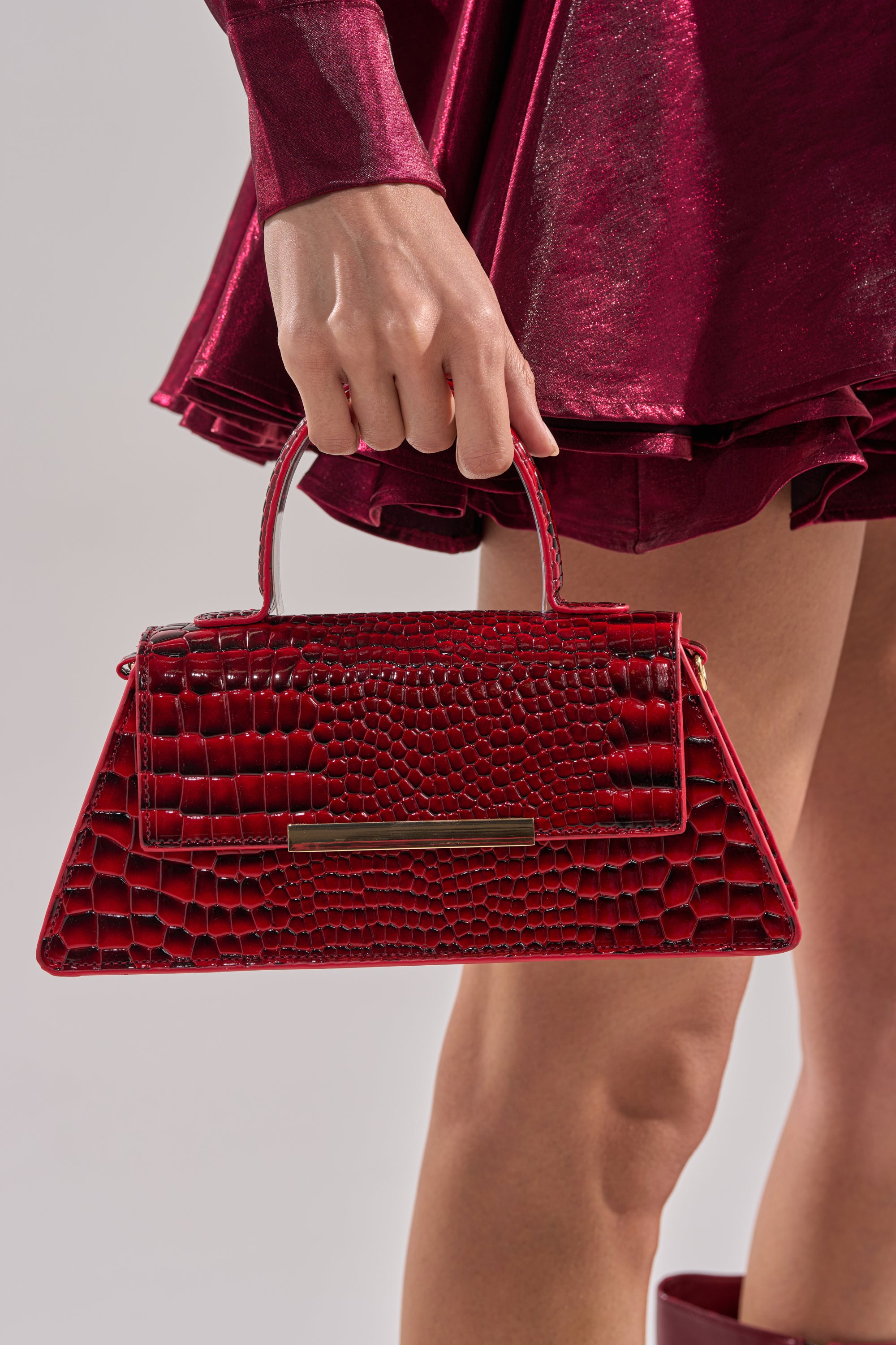 A person in a shiny, dark red dress holds the DAY FAUX CROC PURSE, a cherry red vegan crocodile leather handbag with a short handle and gold rectangular accent, against a plain light background.
