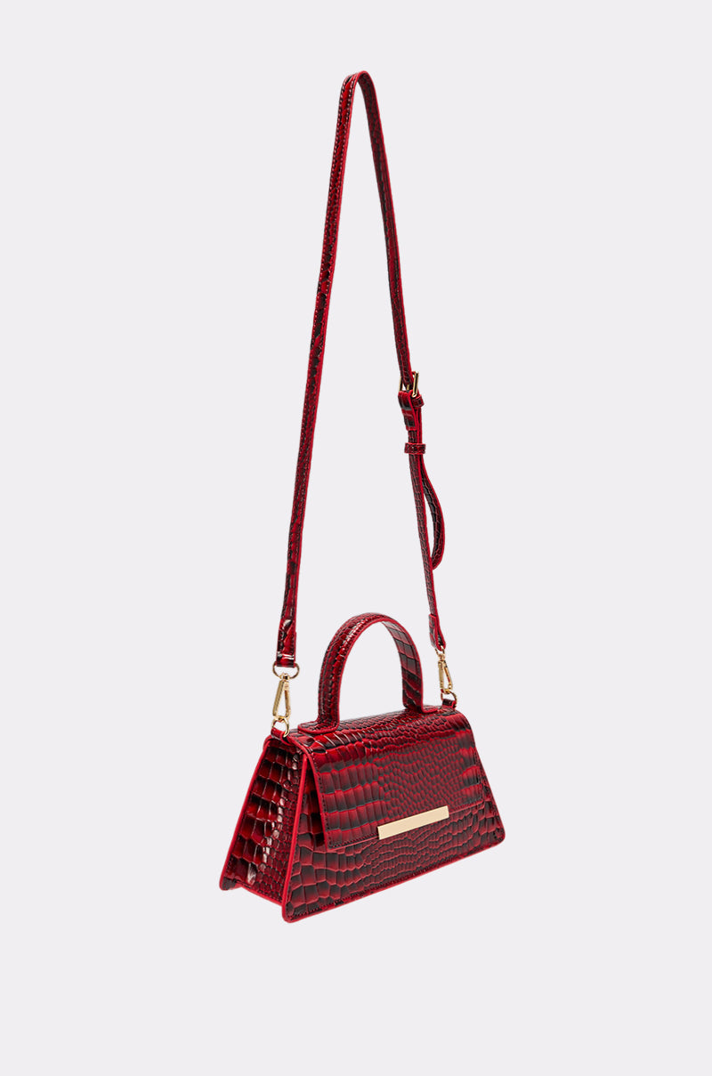 The DAY FAUX CROC PURSE is a small cherry red handbag made from vegan crocodile leather, with a top handle, adjustable shoulder strap, and gold rectangular front plate, shown against a plain light background. 4