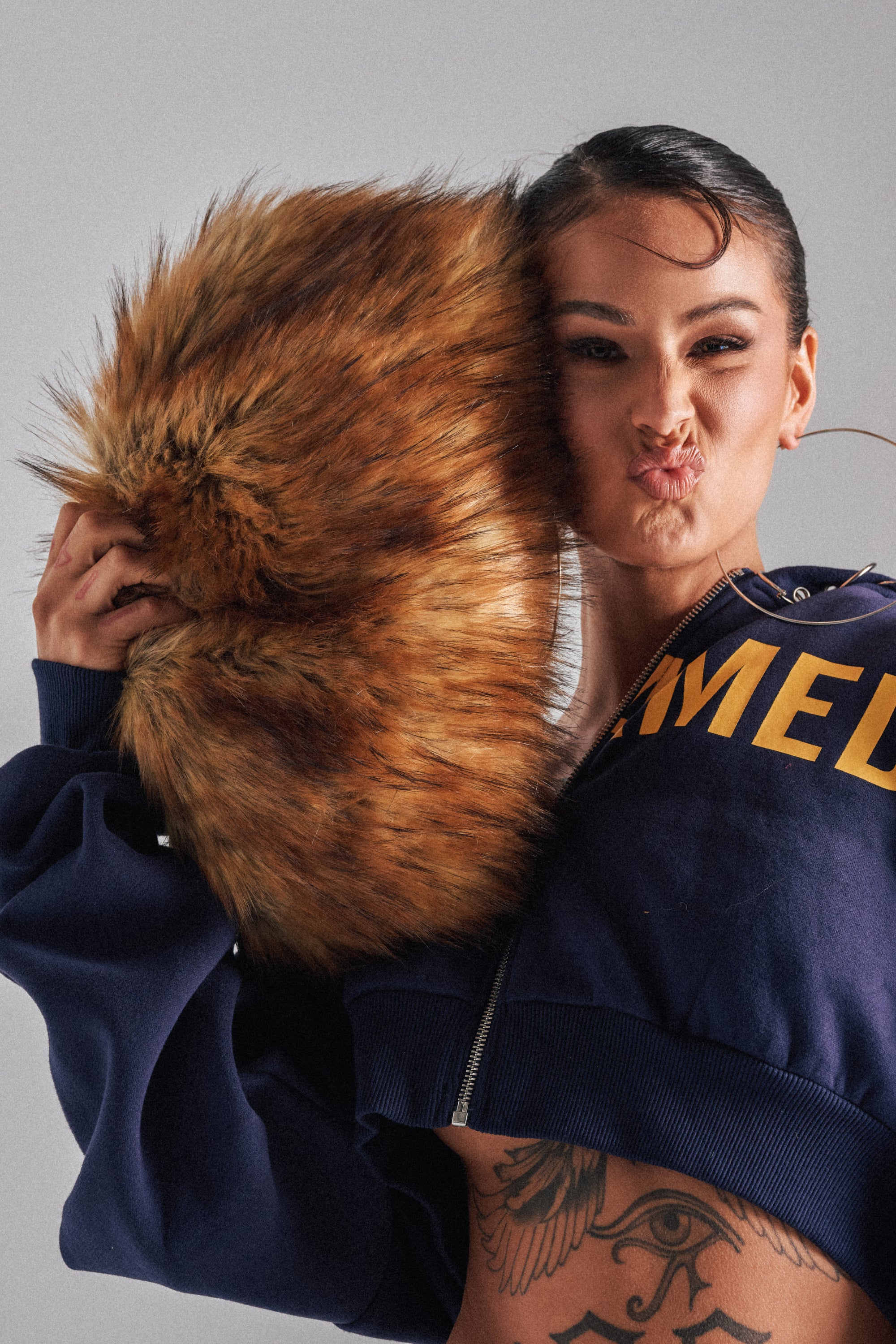 A woman in a navy blue zip-up hoodie playfully holds the WILD CHILD FAUX FUR CLUTCH to her face, with a partial torso tattoo visible. The background is plain gray.