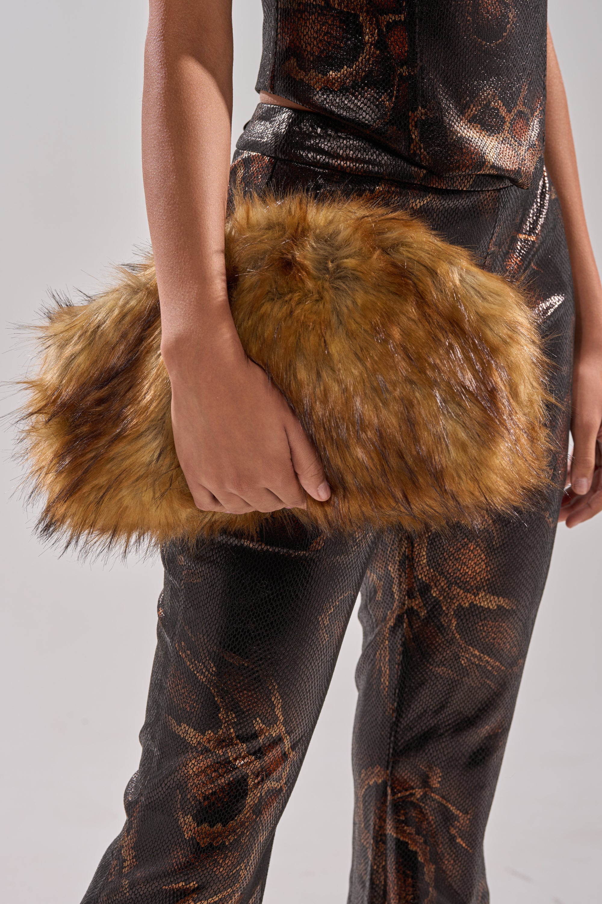 A person in a shiny, black patterned outfit holds the WILD CHILD FAUX FUR CLUTCH under their arm; only their torso, arm, and the clutch are visible in the cropped image.